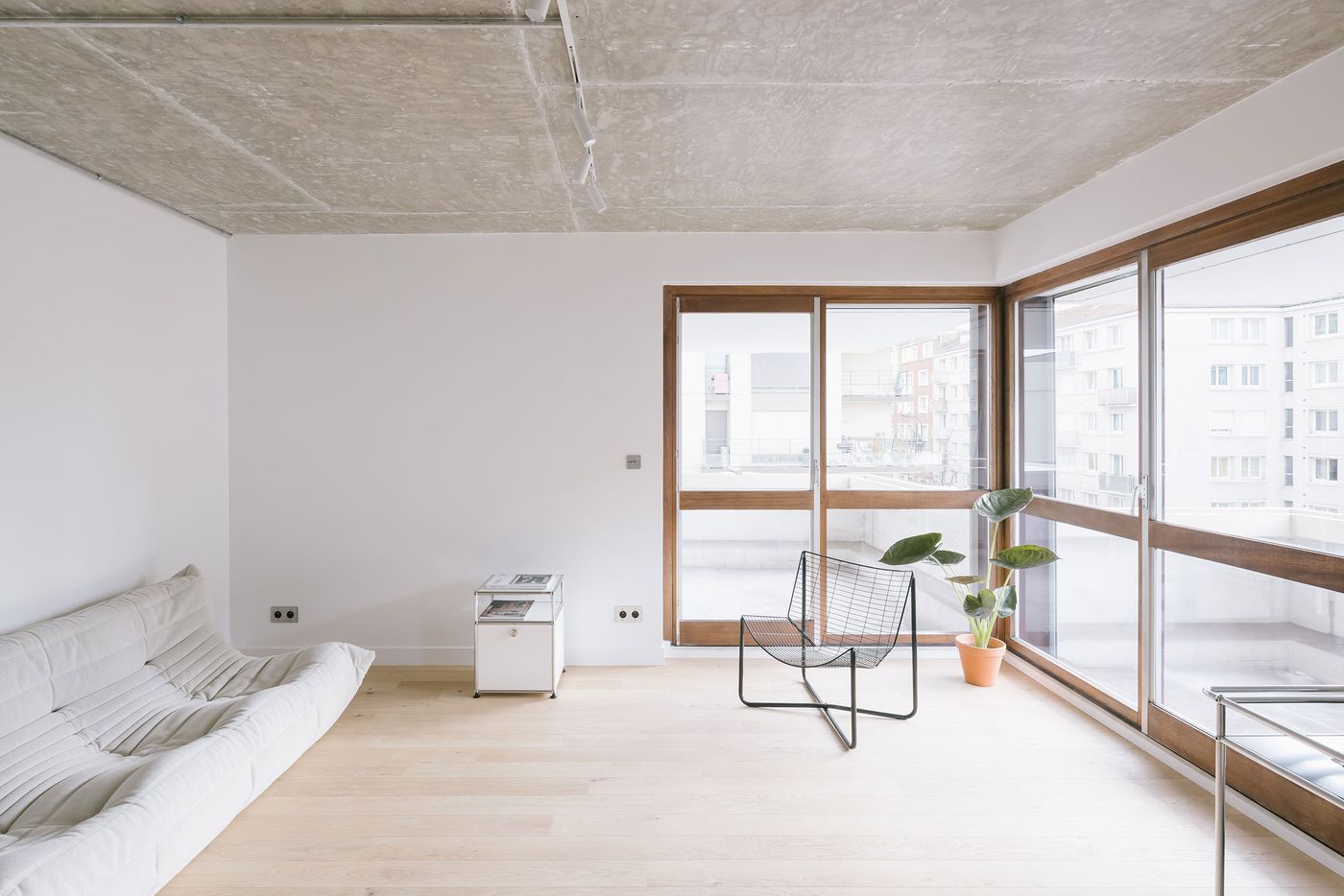 Bright and minimalistic living room with a white sofa, a wire chair, wooden accents, and large windows overlooking an urban view. Bright and minimalistic living room with a white sofa, a wire chair, wooden accents, and large windows overlooking an urban view.