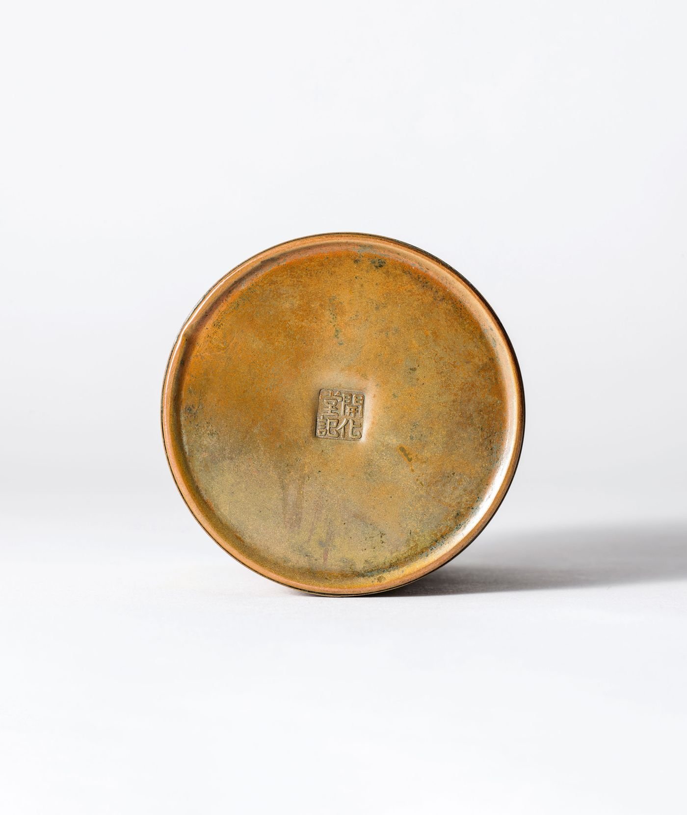 A bronze canister viewed from below, featuring a small engraved square seal in the center and a patina finish. A bronze canister viewed from below, featuring a small engraved square seal in the center and a patina finish.
