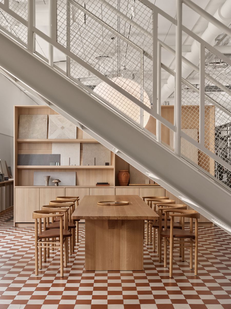 An elegant timber dining table and chairs are positioned under a white industrial staircase with a metal mesh railing. The bold red and white checkerboard floor provides a dynamic contrast to the minimalist wooden furniture and the raw industrial elements above. An elegant timber dining table and chairs are positioned under a white industrial staircase with a metal mesh railing. The bold red and white checkerboard floor provides a dynamic contrast to the minimalist wooden furniture and the raw industrial elements above.