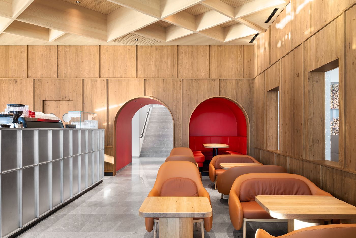 A study in material contrast: the ribbed steel service counter meets warm timber wall cladding beneath a precisely detailed coffered ceiling. Orange leather seating adds tactile warmth to the neutral palette, while the modular ceiling system creates rhythm and visual interest overhead. A study in material contrast: the ribbed steel service counter meets warm timber wall cladding beneath a precisely detailed coffered ceiling. Orange leather seating adds tactile warmth to the neutral palette, while the modular ceiling system creates rhythm and visual interest overhead.