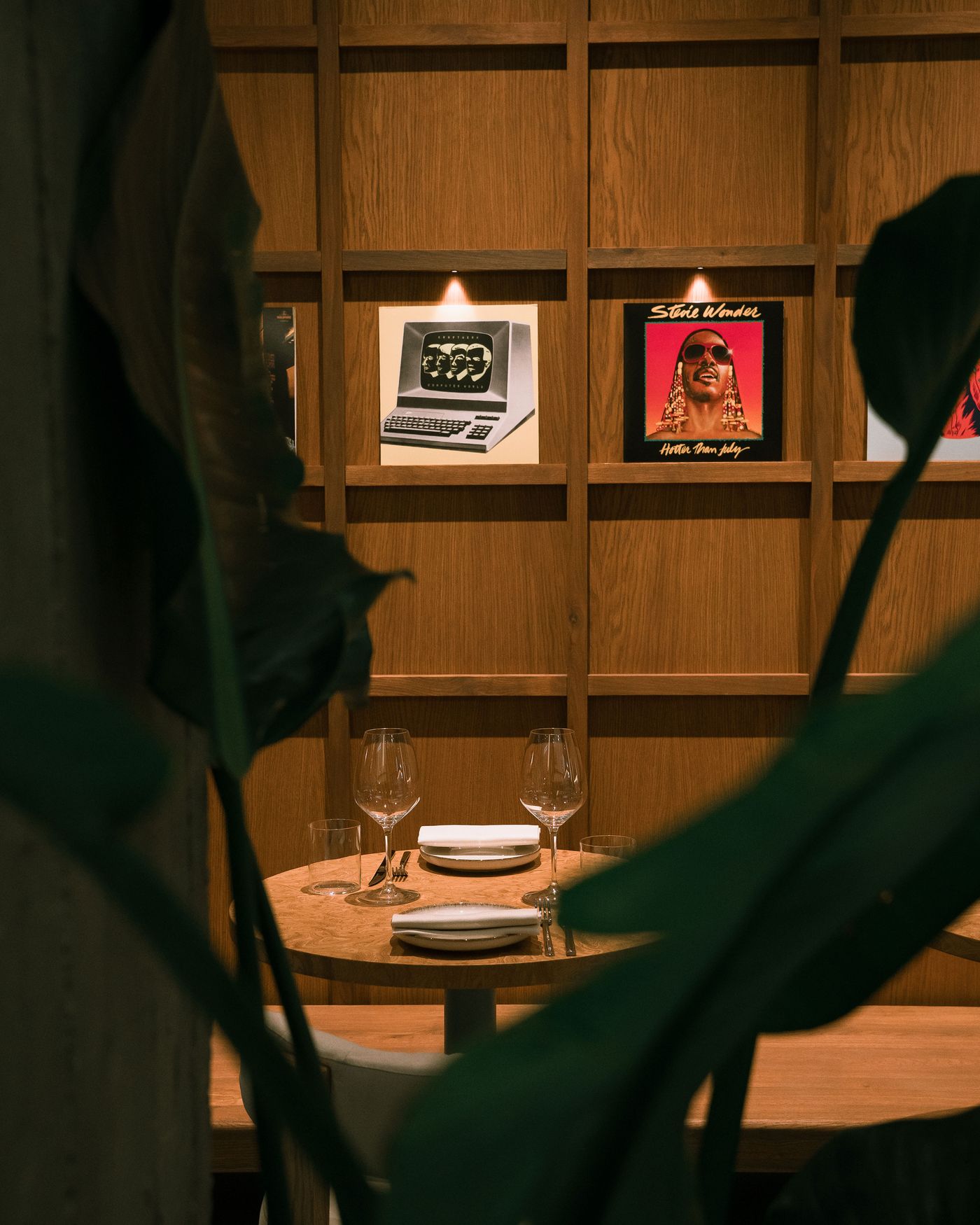 A cozy dining table set for two, surrounded by greenery, with vintage album covers displayed on a wooden wall in the background.