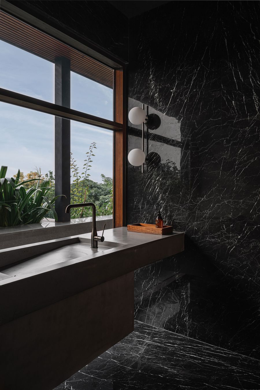 A stylish bathroom features a unique concrete basin with a dark, modern faucet, set against a dramatic black marble wall with prominent white veining. Two spherical wall sconces emit a soft glow. Expansive windows framed in warm wood offer a view of lush greenery and an outdoor concrete wall, creating a serene and sophisticated space that blends natural materials with contemporary design.