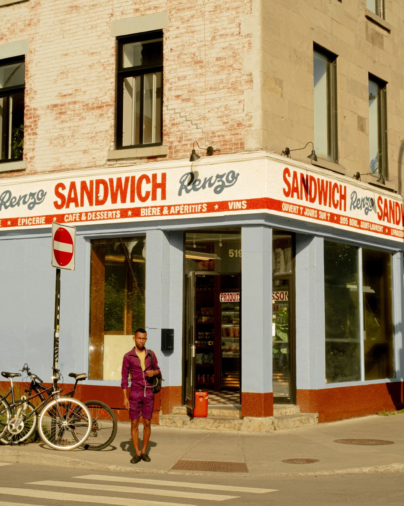 The exterior of Renzo Sandwich shop on a sunny day. The building's facade features a light blue lower level with red trim and signage, and a brick upper level. A young man stands on the sidewalk next to two bicycles. The signage emphasizes "SANDWICH" with the name "Renzo" repeated in a stylized font, indicating a bold, graphic branding.