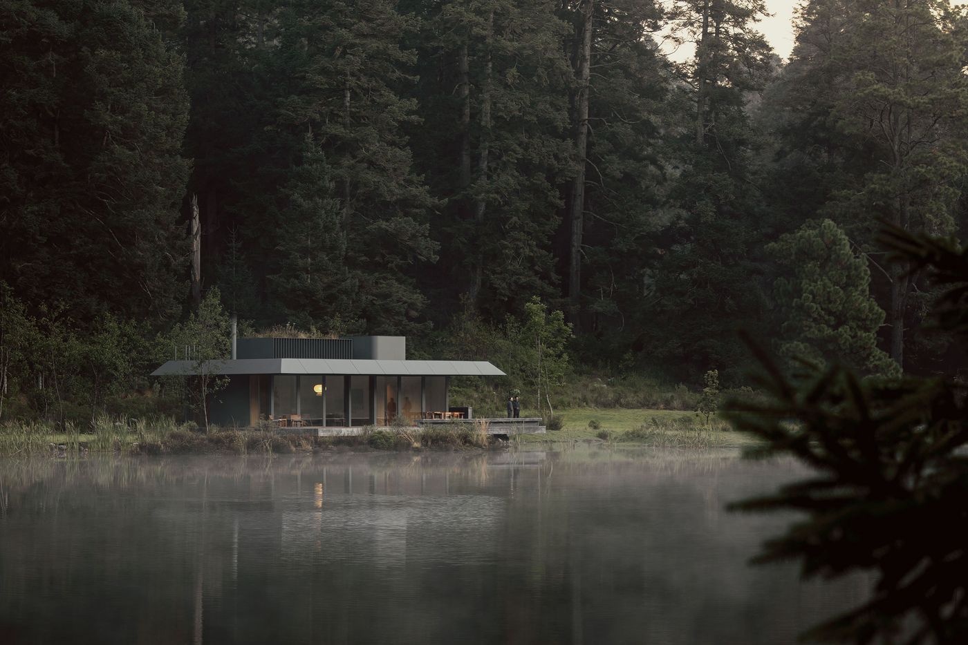A modern, circular cabin with large glass windows sits by a misty lake, surrounded by tall trees and lush greenery.