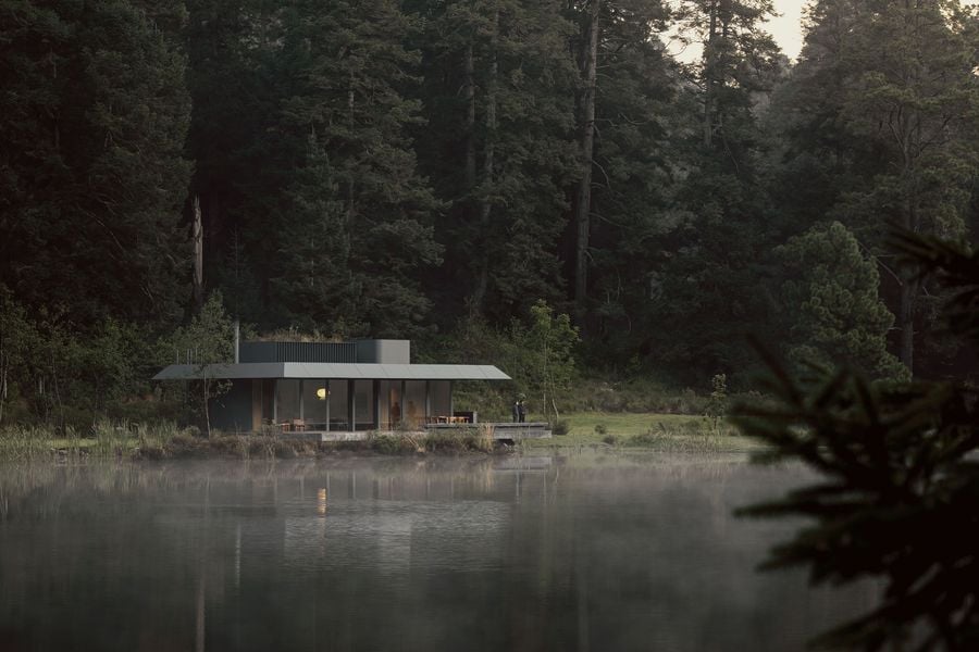 A modern, circular cabin with large glass windows sits by a misty lake, surrounded by tall trees and lush greenery.