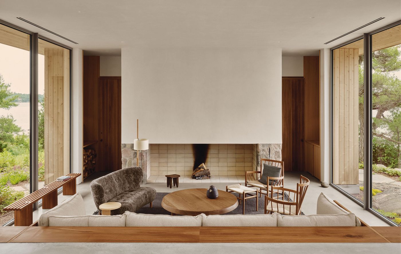 Modern living room with large windows, minimalist furniture, a central round table, and a cozy fireplace surrounded by natural elements. Modern living room with large windows, minimalist furniture, a central round table, and a cozy fireplace surrounded by natural elements.