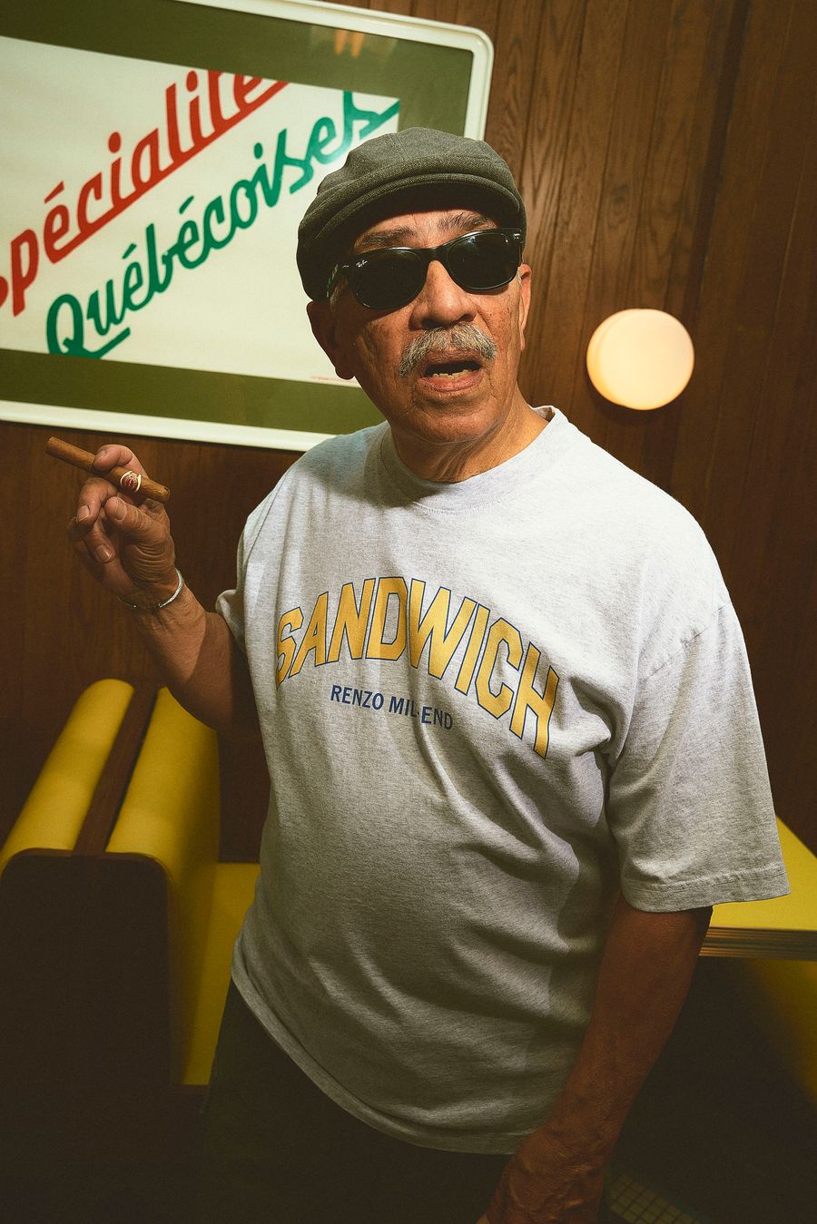 A man in a flat cap and sunglasses poses, holding a cigar. He wears a light grey t-shirt with "SANDWICH" in bold yellow lettering, beneath which is "RENZO MILANO." The background shows wood paneling and a yellow booth, consistent with a vintage diner aesthetic.