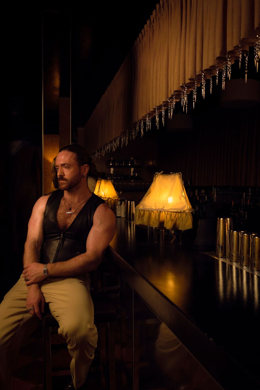 A man with a contemplative expression, wearing a black leather vest, leans casually on a dark, polished bar at The Roses of Elagabalus. Behind him, vintage-inspired lamps with ruffled shades emit a warm, inviting glow, complemented by the unique, cascading icicle-like embellishments suspended above the bar, contributing to the sultry, retro mood. A man with a contemplative expression, wearing a black leather vest, leans casually on a dark, polished bar at The Roses of Elagabalus. Behind him, vintage-inspired lamps with ruffled shades emit a warm, inviting glow, complemented by the unique, cascading icicle-like embellishments suspended above the bar, contributing to the sultry, retro mood.