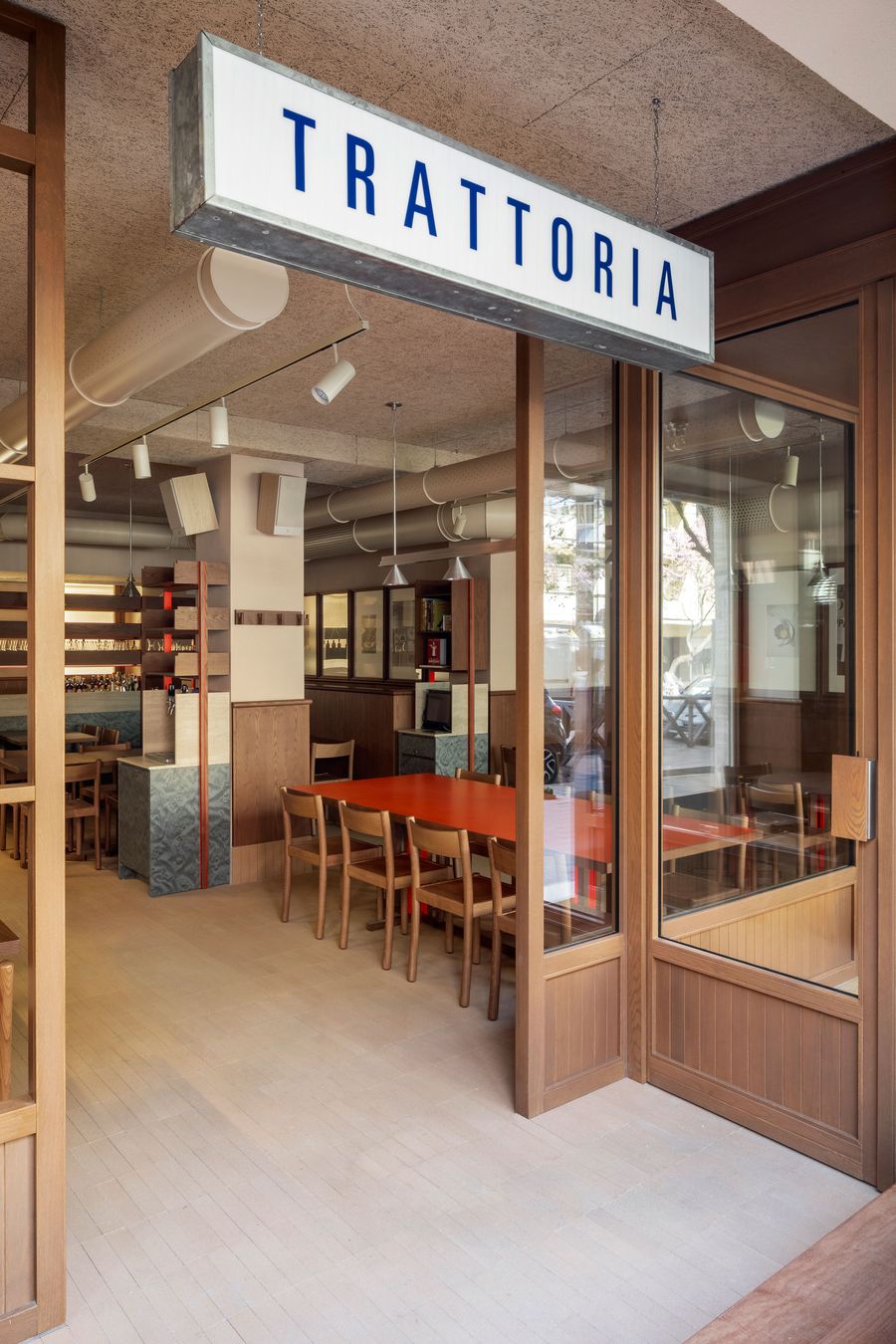 The entrance to Santo Palato in Rome, designed by Naessi Studio, under a prominent "TRATTORIA" sign. Large wooden-framed glass doors open into a bright, inviting space. Inside, a vibrant orange communal table is visible, surrounded by minimalist wooden chairs, hinting at the modern yet authentic dining experience within. The entrance to Santo Palato in Rome, designed by Naessi Studio, under a prominent "TRATTORIA" sign. Large wooden-framed glass doors open into a bright, inviting space. Inside, a vibrant orange communal table is visible, surrounded by minimalist wooden chairs, hinting at the modern yet authentic dining experience within.