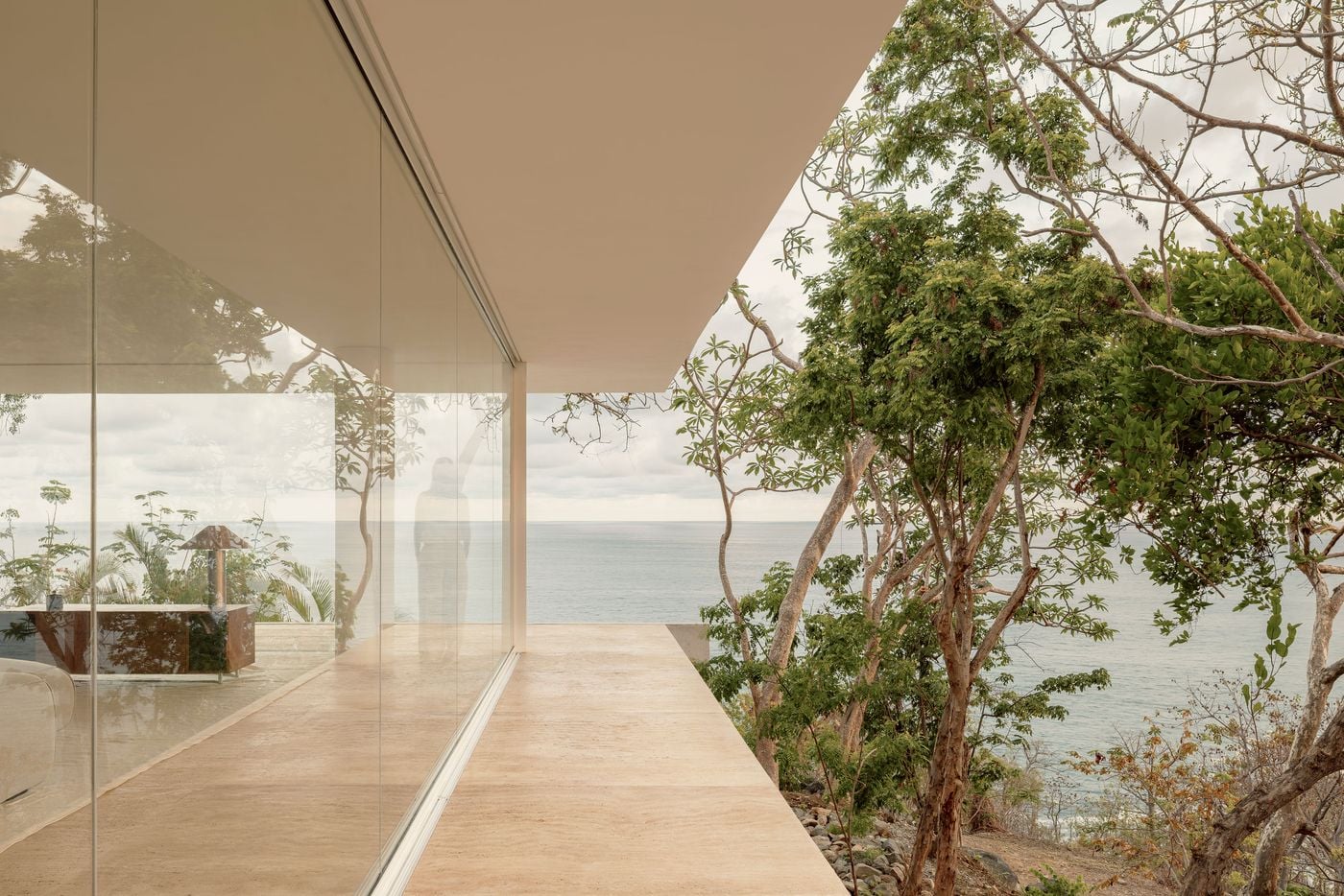 At a corner of the living level, slim-framed glazing doubles as a mirror, layering reflected branches and a faint silhouette over a timber sideboard and faceted table lamp inside. The pale terrace ends in a clean, unguarded edge, with twisting trunks and the open sea filling the view beyond. At a corner of the living level, slim-framed glazing doubles as a mirror, layering reflected branches and a faint silhouette over a timber sideboard and faceted table lamp inside. The pale terrace ends in a clean, unguarded edge, with twisting trunks and the open sea filling the view beyond.