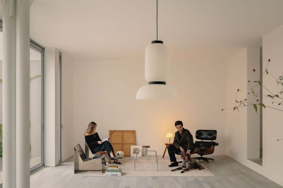 Casa Tlaloc’s minimalist living room pairs white walls and pale grey stone flooring with a carefully edited mix of sculptural metal seating, warm timber accents and black leather. A large paper pendant anchors the otherwise spare composition as two occupants and their dog bring an informal domestic presence to the space.