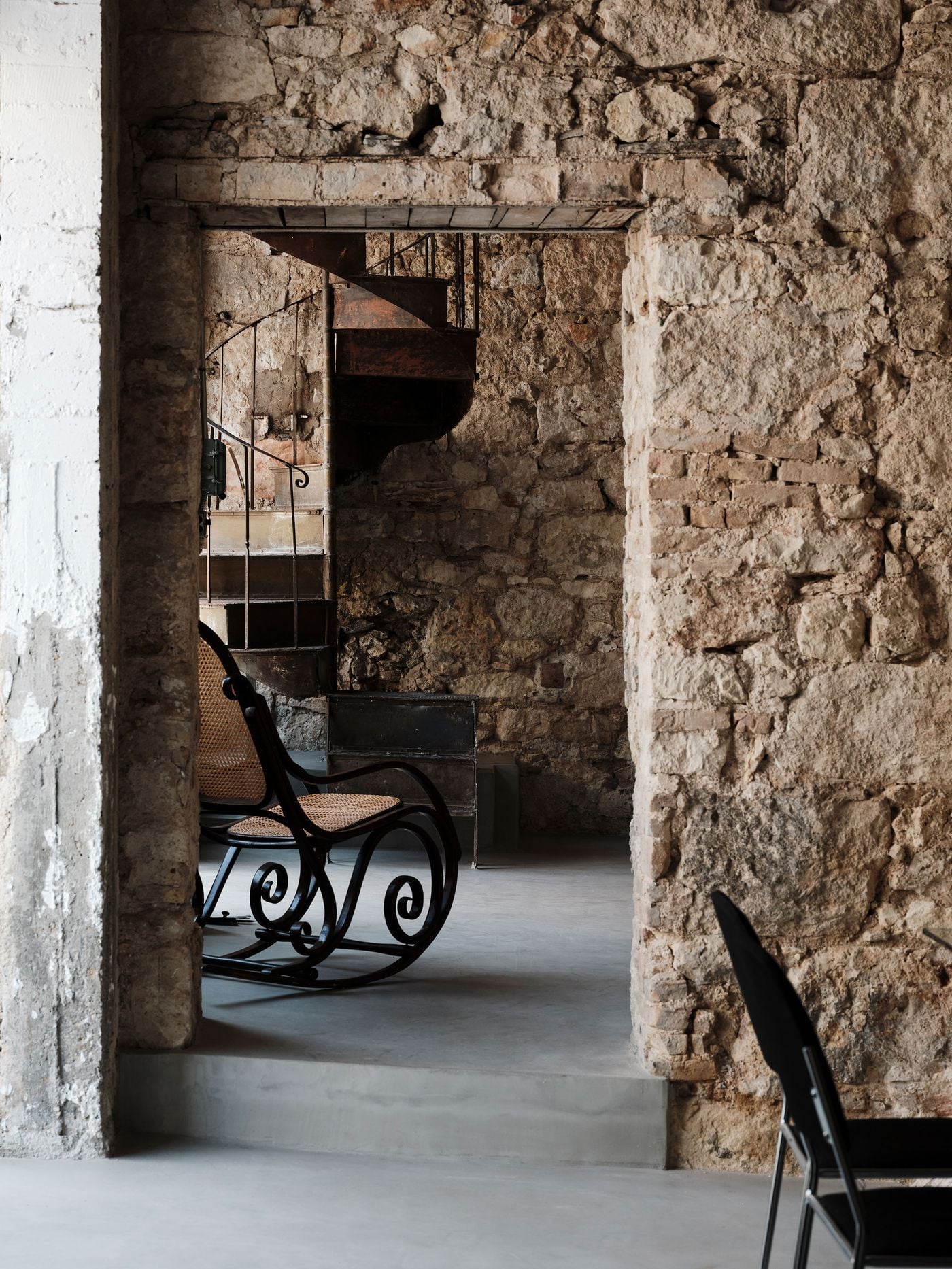 Viewed through a thick masonry opening, a vintage cane rocking chair sits against exposed stone walls beside the rusted spiral staircase. The composition distils MPNYC’s palimpsest approach: fragments belonging to different eras coexist without hierarchy, unified by the restrained grey flooring and the warm, weathered tones of the original fabric. Viewed through a thick masonry opening, a vintage cane rocking chair sits against exposed stone walls beside the rusted spiral staircase. The composition distils MPNYC’s palimpsest approach: fragments belonging to different eras coexist without hierarchy, unified by the restrained grey flooring and the warm, weathered tones of the original fabric.