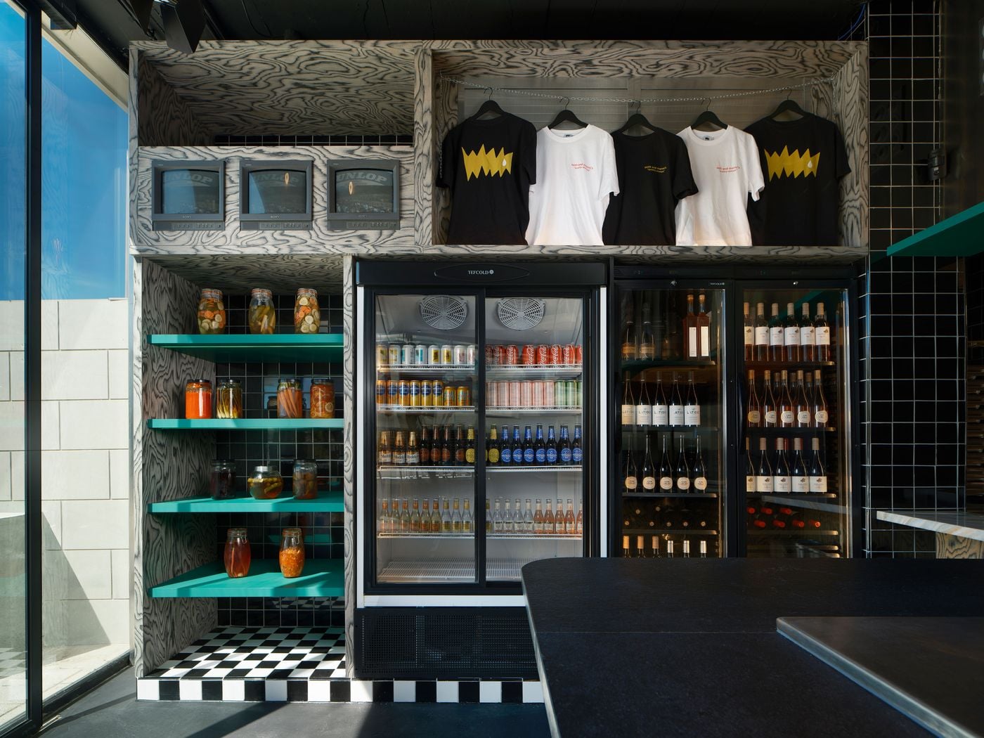 A deli corner at Seth & Manni's combines grey wood-grain cabinetry, turquoise shelves of pickle jars and glass-fronted fridges stocked with beer and wine. Overhead, branded T-shirts hang from a chain beside a row of vintage Sony monitors. A black-and-white checkerboard floor grounds the display. A deli corner at Seth & Manni's combines grey wood-grain cabinetry, turquoise shelves of pickle jars and glass-fronted fridges stocked with beer and wine. Overhead, branded T-shirts hang from a chain beside a row of vintage Sony monitors. A black-and-white checkerboard floor grounds the display.