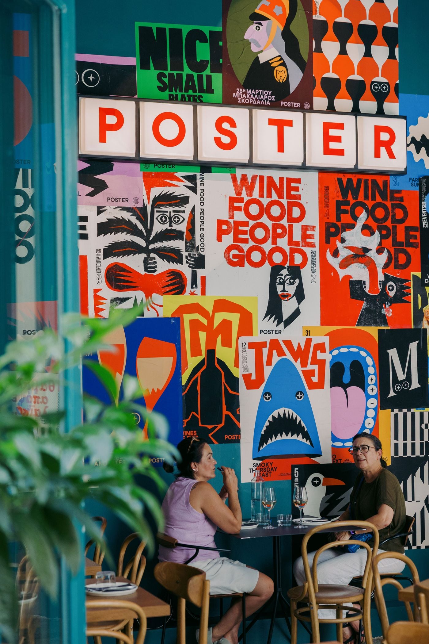 Poster restaurant in Thessaloniki: two diners sit beneath a maximalist wall of bold screen-printed posters in red, blue and yellow, including a shark graphic, with an illuminated POSTER marquee sign, teal walls and bentwood chairs.