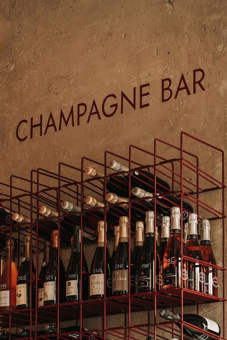 An exposed concrete column bearing the words ‘CHAMPAGNE BAR’ provides a rugged backdrop for WIN's slender burgundy metal wine racks. The open-grid shelving accommodates bottles in varying orientations, creating a disciplined yet visually dynamic display that reflects the project's interplay between industrial rigour and the organic character of wine production. An exposed concrete column bearing the words ‘CHAMPAGNE BAR’ provides a rugged backdrop for WIN's slender burgundy metal wine racks. The open-grid shelving accommodates bottles in varying orientations, creating a disciplined yet visually dynamic display that reflects the project's interplay between industrial rigour and the organic character of wine production.
