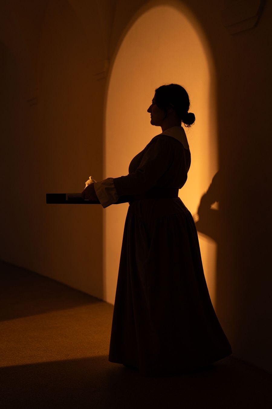 A server in a long, full-skirted dress with a white collar is caught in silhouette at Castel Badia, carrying a tray with a lidded porcelain pot. Behind her, a tall arched opening glows amber against the darkened vaulted interior, giving the scene the stillness of a painted tableau. A server in a long, full-skirted dress with a white collar is caught in silhouette at Castel Badia, carrying a tray with a lidded porcelain pot. Behind her, a tall arched opening glows amber against the darkened vaulted interior, giving the scene the stillness of a painted tableau.