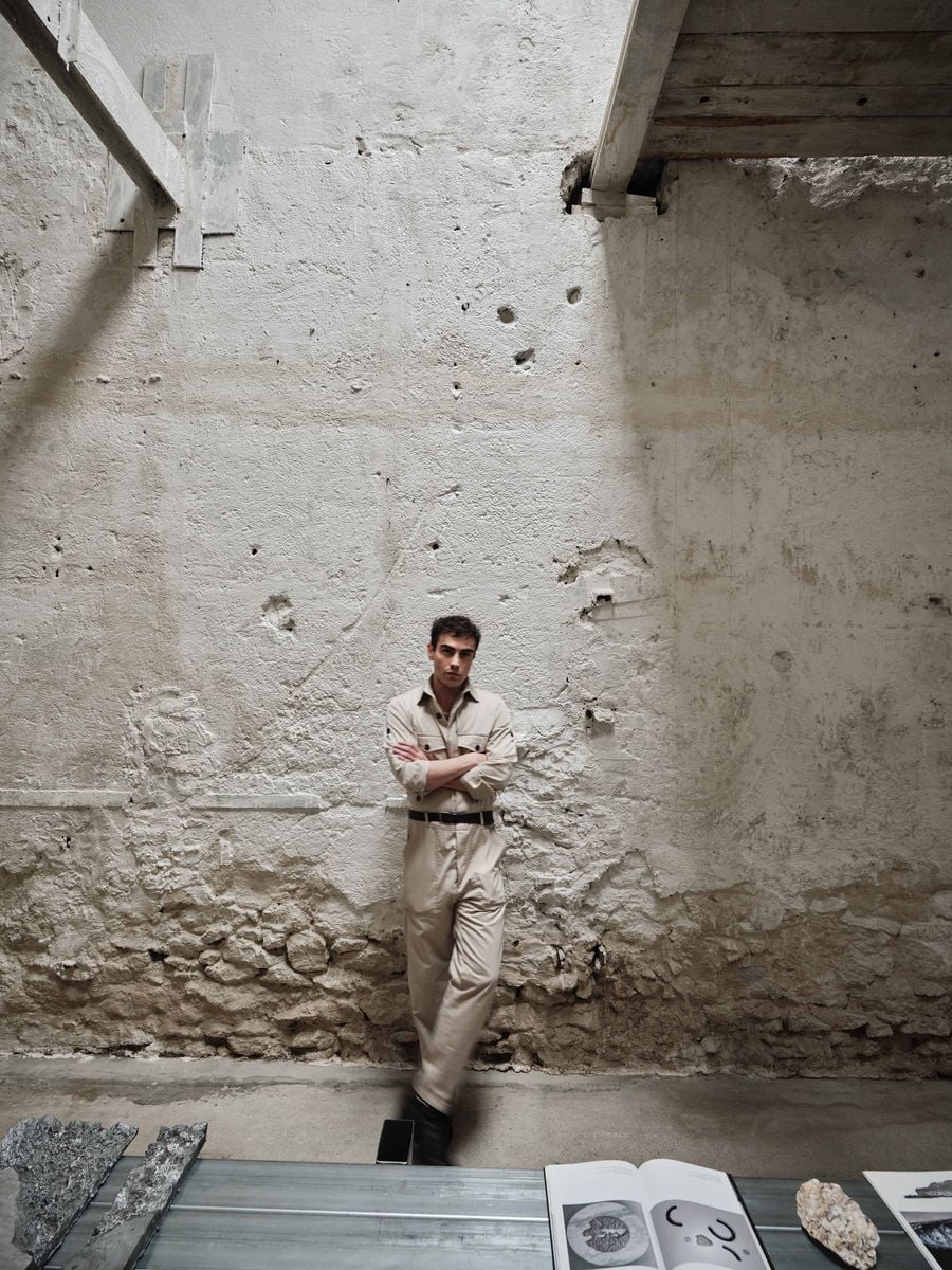 Architect Andreas Kostopoulos stands against the weathered masonry of Castor Place, giving a sense of scale to the former warehouse’s tall, time-worn interior. In the foreground, a slender metal display table holds publications and material samples, while daylight from above picks out the wall’s accumulated layers of stone, plaster and structural traces. Architect Andreas Kostopoulos stands against the weathered masonry of Castor Place, giving a sense of scale to the former warehouse’s tall, time-worn interior. In the foreground, a slender metal display table holds publications and material samples, while daylight from above picks out the wall’s accumulated layers of stone, plaster and structural traces.