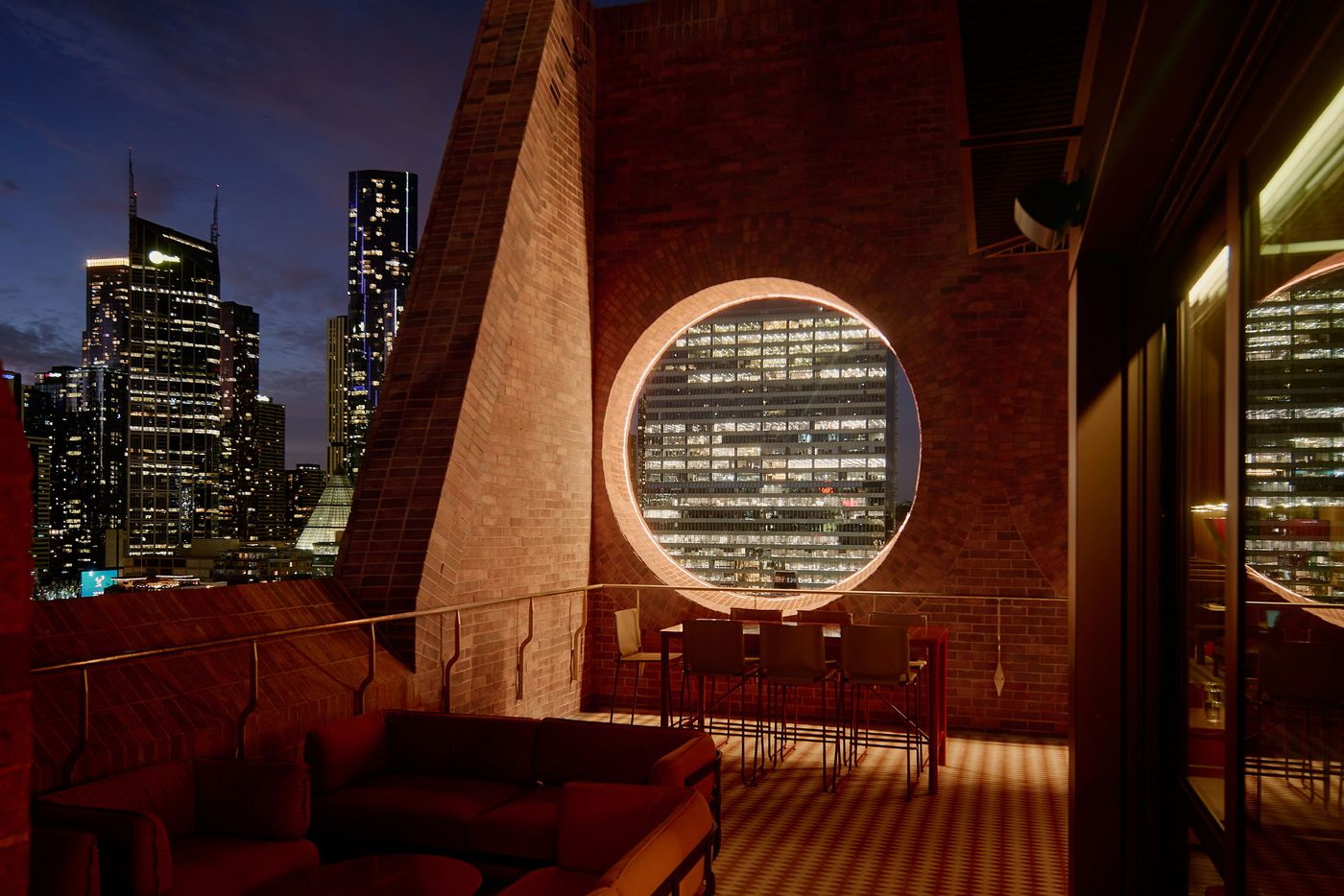 A rooftop lounge with modern seating overlooks a vibrant city skyline illuminated at night through a circular window.