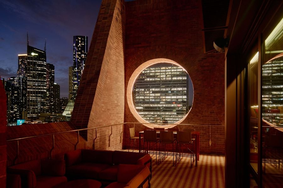 A rooftop lounge with modern seating overlooks a vibrant city skyline illuminated at night through a circular window.