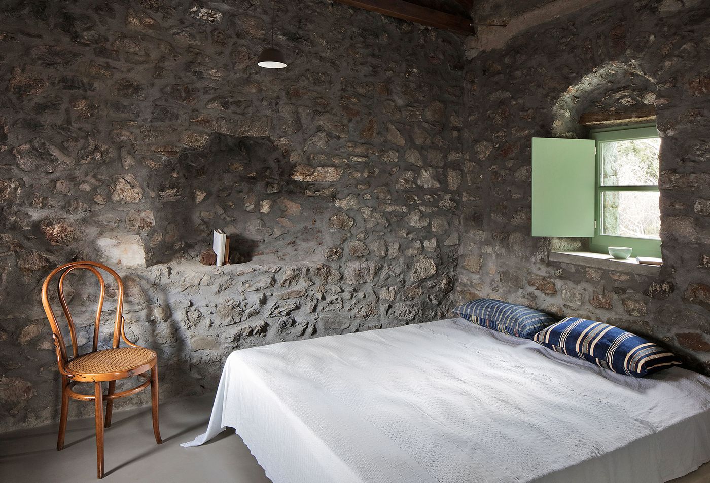 Monastic bedroom celebrates the building's contemplative character through minimal furnishing and raw stone walls, where sage-green window frames provide subtle contemporary detail. The traditional bentwood chair and simple bedding demonstrate how authentic Mediterranean living can be maintained while incorporating necessary modern comfort within the preserved architectural shell.