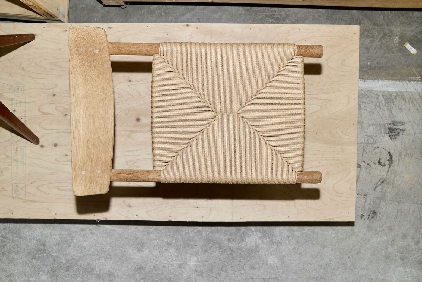 An overhead view of a wooden chair with a woven natural fiber seat, resting on a plywood surface.