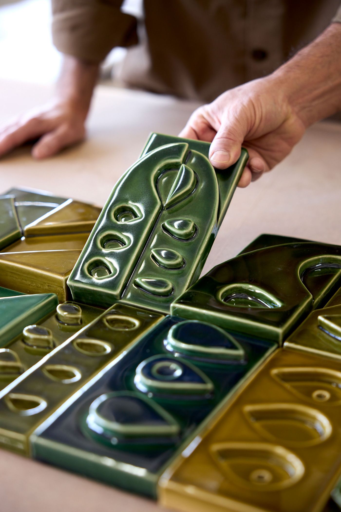 A craftsman's hands carefully arrange an exquisite green glazed ceramic tile, part of the "Arcadia" collection by Oficina Marques and Viúva Lamego. The tile features an elegant, elongated teardrop motif with circular accents. Surrounding tiles in varying shades of green and amber reveal the intricate, modular design, highlighting the artisanal quality. A craftsman's hands carefully arrange an exquisite green glazed ceramic tile, part of the "Arcadia" collection by Oficina Marques and Viúva Lamego. The tile features an elegant, elongated teardrop motif with circular accents. Surrounding tiles in varying shades of green and amber reveal the intricate, modular design, highlighting the artisanal quality.