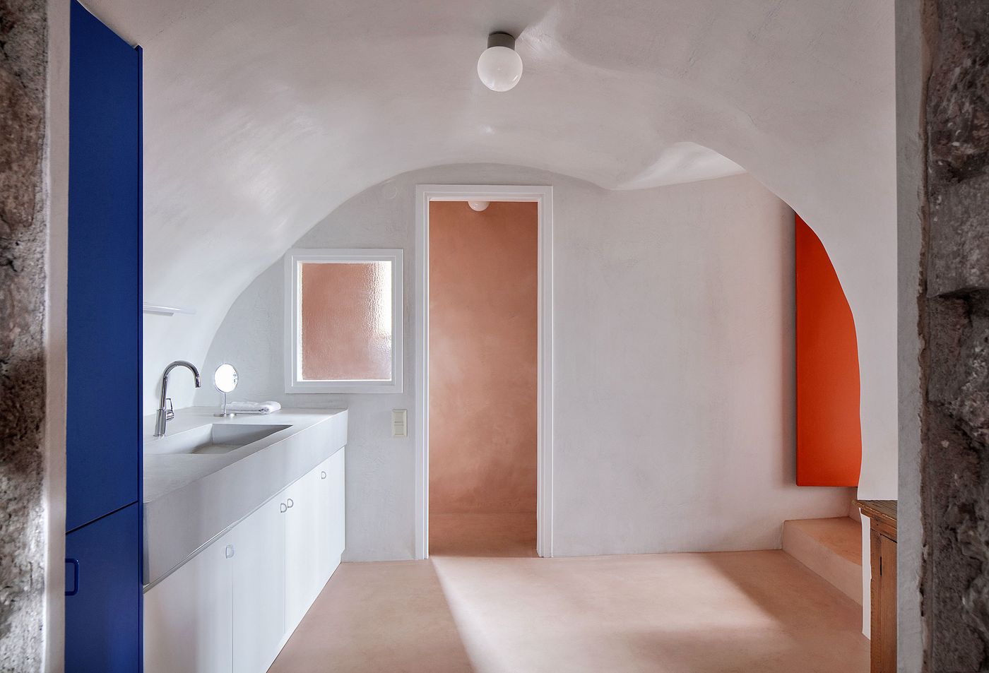 Contemporary bathroom pod floats within the ancient stone shell, where bold coral and blue color blocks create striking contrast against the whitewashed barrel vault. The minimalist intervention demonstrates how modern amenities can be inserted into historical spaces while maintaining the building's authentic architectural character through careful spatial choreography.