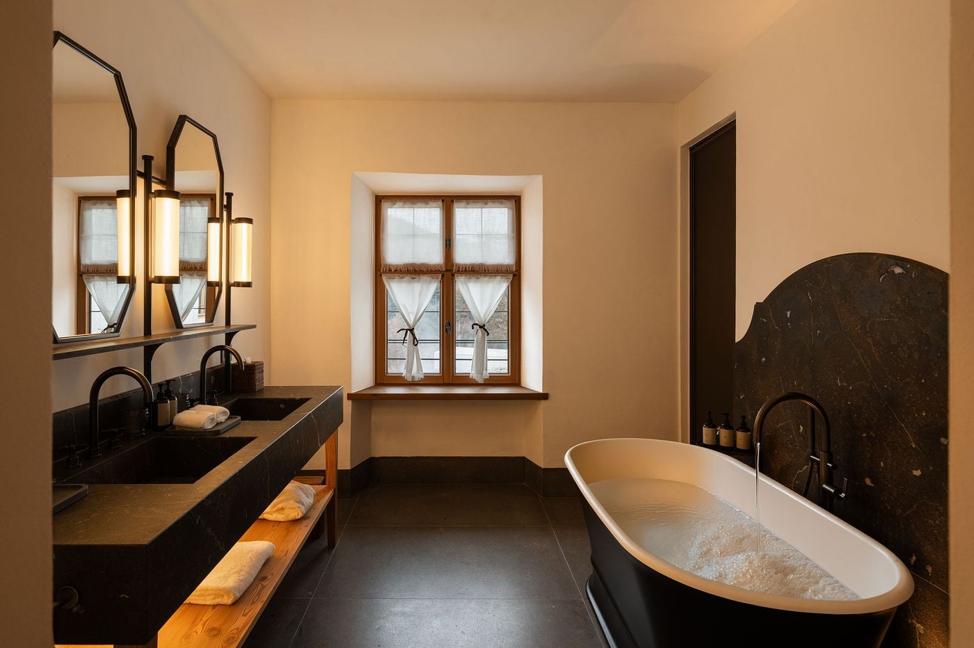A bathroom at Castel Badia pairs a monolithic dark stone double vanity, elongated octagonal mirrors and cylindrical sconces with a pine shelf below. A freestanding bathtub fills before a shaped dark stone splashback, while linen café curtains tied with ribbons soften the deep-set timber window. A bathroom at Castel Badia pairs a monolithic dark stone double vanity, elongated octagonal mirrors and cylindrical sconces with a pine shelf below. A freestanding bathtub fills before a shaped dark stone splashback, while linen café curtains tied with ribbons soften the deep-set timber window.