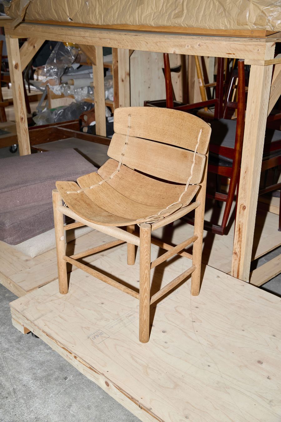 A wooden chair with a curved, slatted seat and backrest, displayed on a wooden platform among other furniture pieces in a storage area. A wooden chair with a curved, slatted seat and backrest, displayed on a wooden platform among other furniture pieces in a storage area.