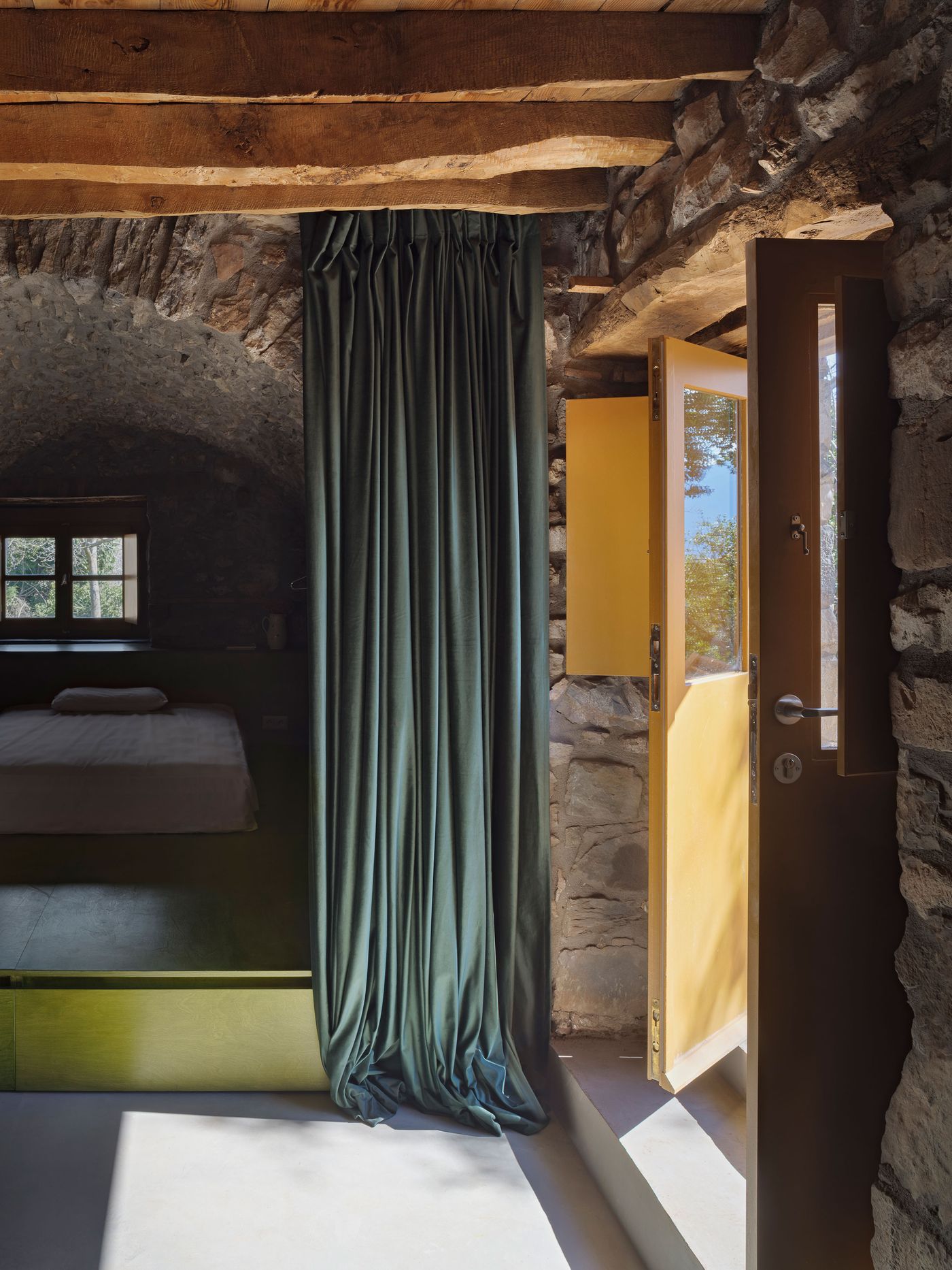 Dramatic emerald-green curtains create theatrical privacy within the exposed stone interior, where yellow contemporary elements provide cheerful accent against the raw masonry walls. The composition reveals the project's layered approach to restoration, celebrating both the building's rugged authenticity and contemporary comfort through bold color interventions.