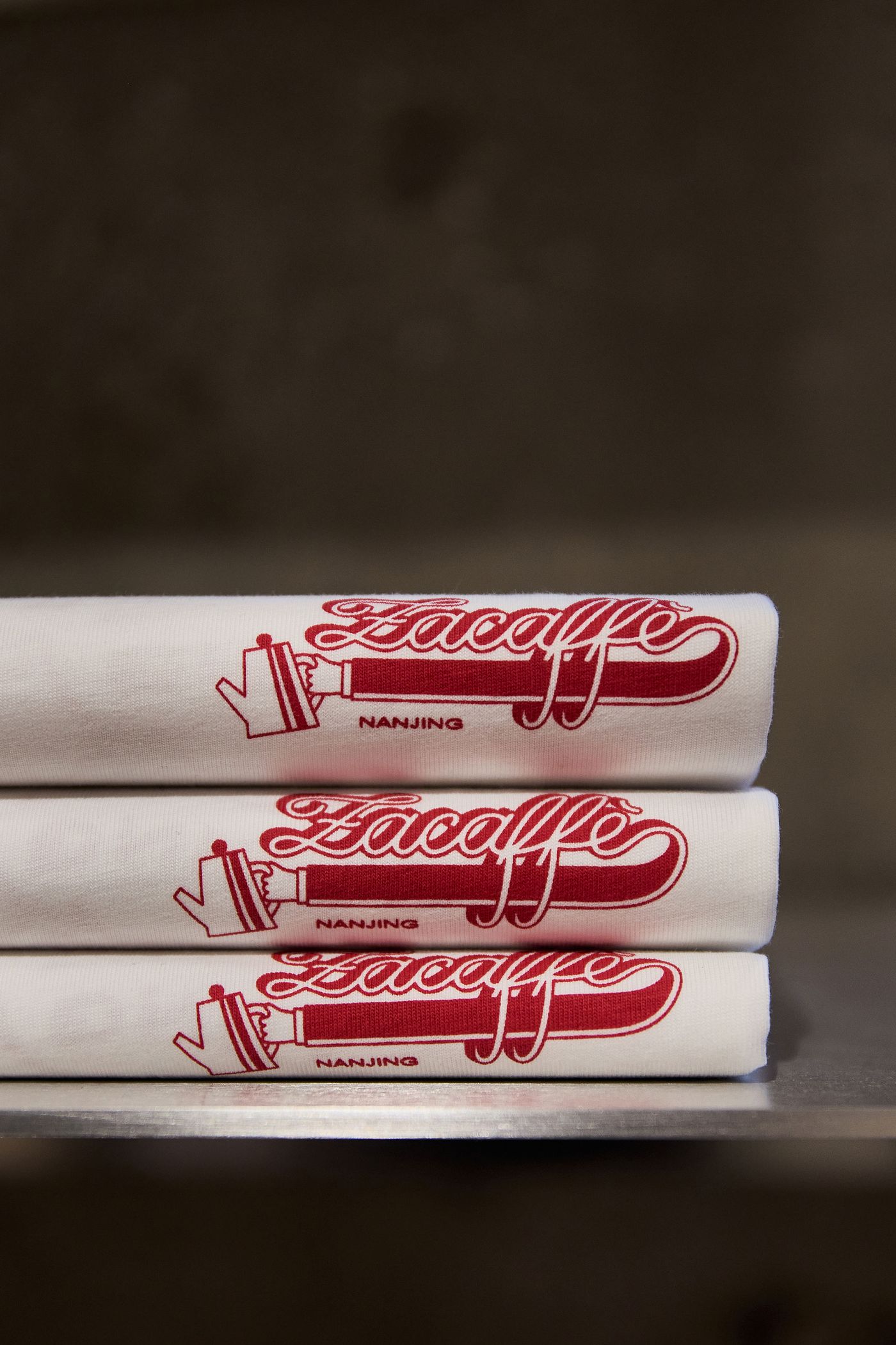 Three neatly stacked white t-shirts, each displaying a red "Zacaffè" logo and design, labeled "Nanjing." Three neatly stacked white t-shirts, each displaying a red "Zacaffè" logo and design, labeled "Nanjing."