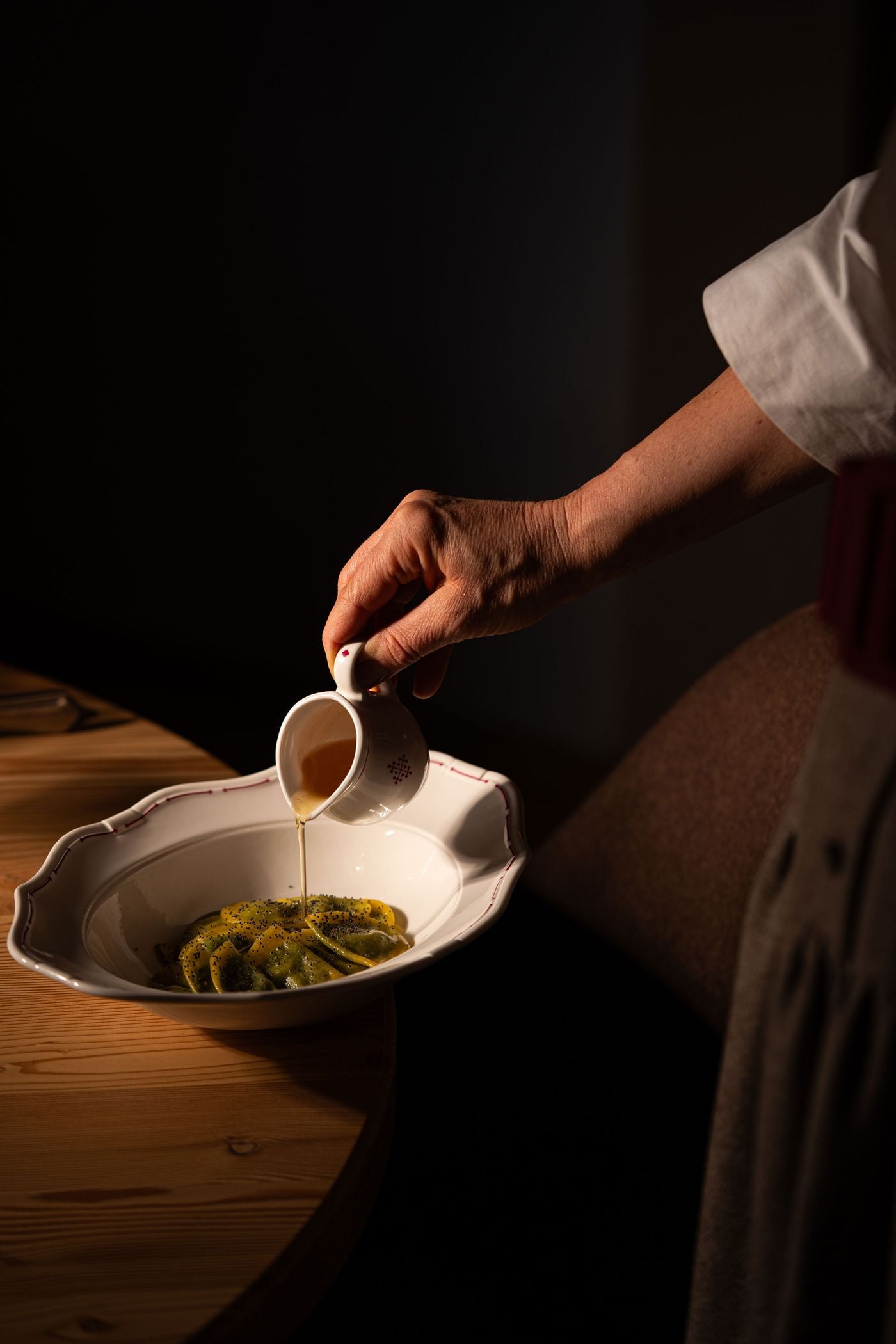 A hand pours golden melted butter from a small jug bearing the hotel's cross emblem over half-moon pasta sprinkled with poppy seeds, a dish resembling South Tyrolean Schlutzkrapfen. The scalloped porcelain bowl, edged in red, rests on a pine table against a deep, painterly darkness. A hand pours golden melted butter from a small jug bearing the hotel's cross emblem over half-moon pasta sprinkled with poppy seeds, a dish resembling South Tyrolean Schlutzkrapfen. The scalloped porcelain bowl, edged in red, rests on a pine table against a deep, painterly darkness.