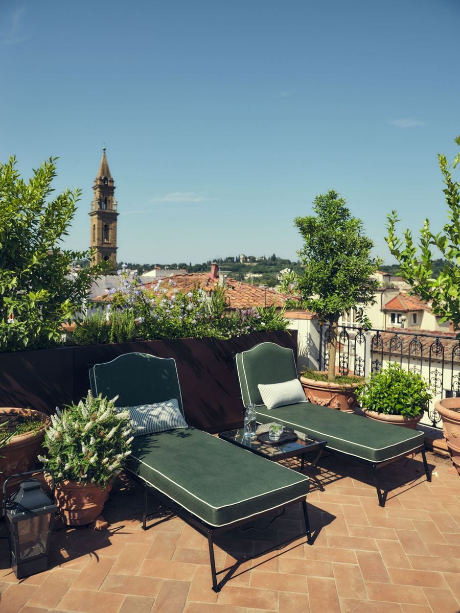 Two green chaise longues with white piping sit on herringbone terracotta on the rooftop terrace of La Réserve Firenze, flanked by citrus trees in garlanded terracotta urns. Beyond a planted parapet and iron railing, a stone bell tower rises against a clear sky above a patchwork of tiled roofs and wooded hills. Two green chaise longues with white piping sit on herringbone terracotta on the rooftop terrace of La Réserve Firenze, flanked by citrus trees in garlanded terracotta urns. Beyond a planted parapet and iron railing, a stone bell tower rises against a clear sky above a patchwork of tiled roofs and wooded hills.