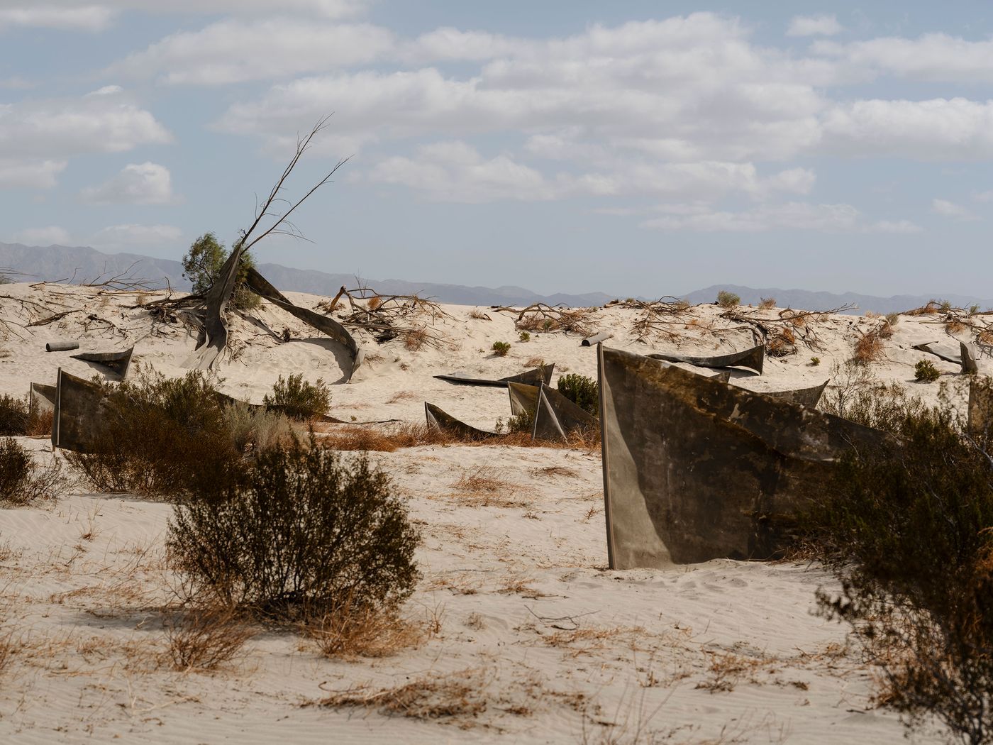 Desolate landscape with abandoned, weathered boats half-buried in sand, surrounded by sparse vegetation and distant mountains.