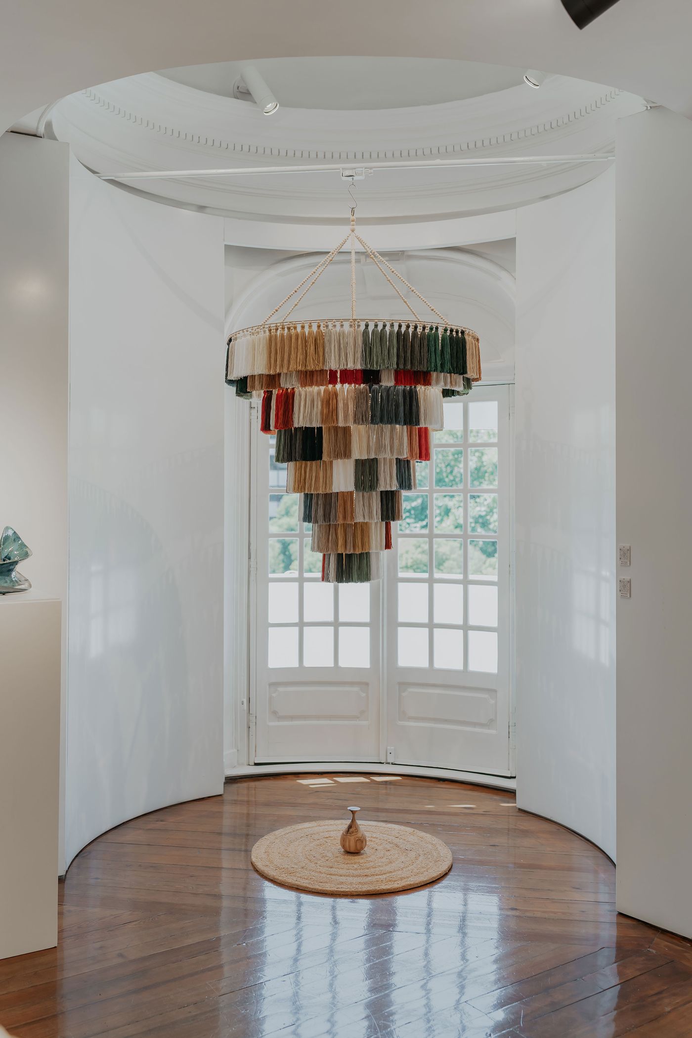 A grand, multi-tiered chandelier crafted from a multitude of colored tassels hangs prominently in the Roca Lisboa Gallery's "Echo Raw Materials" exhibition. Below, a small, elegant vase sits on a round woven mat, centered on a polished wooden floor. The semi-circular white walls and large French doors create a dramatic, light-filled backdrop for this artistic installation. A grand, multi-tiered chandelier crafted from a multitude of colored tassels hangs prominently in the Roca Lisboa Gallery's "Echo Raw Materials" exhibition. Below, a small, elegant vase sits on a round woven mat, centered on a polished wooden floor. The semi-circular white walls and large French doors create a dramatic, light-filled backdrop for this artistic installation.