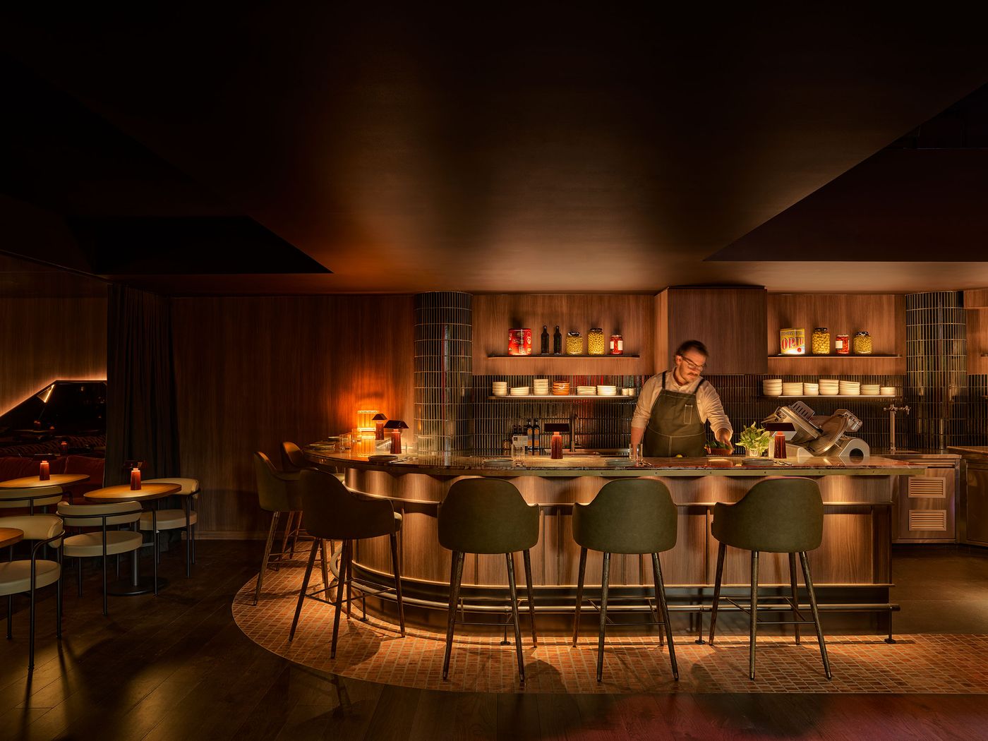 A dimly lit, stylish bar with wooden accents, elegant seating, and shelves lined with bottles and jars behind the countertop.
