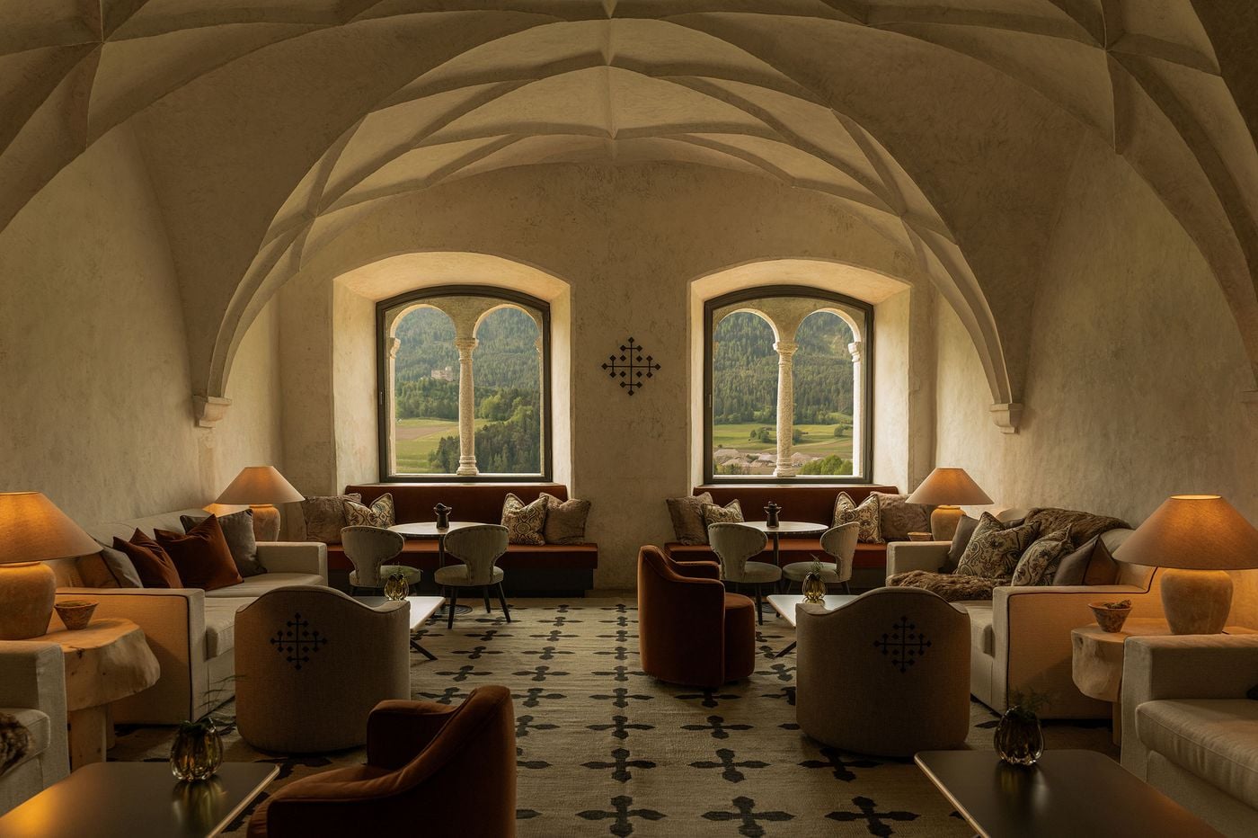 In summer, the former Abbess's Hall at Castel Badia sits beneath its Gothic net vault, its twin-arched stone windows framing green meadows and a distant hilltop castle. A rug patterned with the hotel's cross emblem anchors cream sofas, cross-embroidered tub chairs, burnt-orange velvet armchairs and terracotta lamps in a symmetrical arrangement. In summer, the former Abbess's Hall at Castel Badia sits beneath its Gothic net vault, its twin-arched stone windows framing green meadows and a distant hilltop castle. A rug patterned with the hotel's cross emblem anchors cream sofas, cross-embroidered tub chairs, burnt-orange velvet armchairs and terracotta lamps in a symmetrical arrangement.