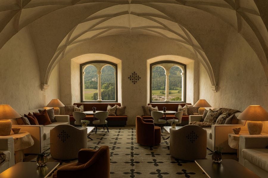 In summer, the former Abbess's Hall at Castel Badia sits beneath its Gothic net vault, its twin-arched stone windows framing green meadows and a distant hilltop castle. A rug patterned with the hotel's cross emblem anchors cream sofas, cross-embroidered tub chairs, burnt-orange velvet armchairs and terracotta lamps in a symmetrical arrangement.