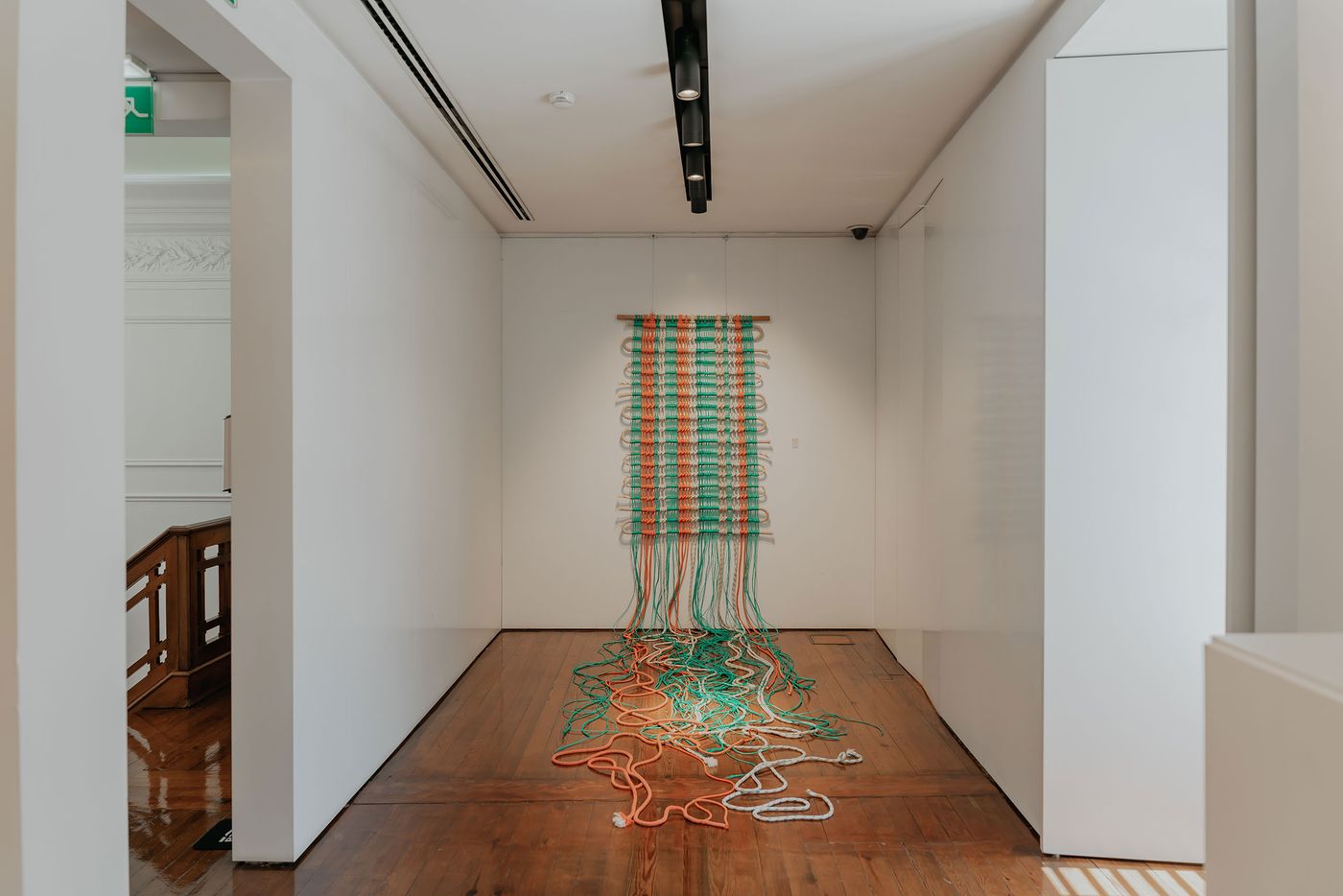An innovative textile installation, part of the "Echo Raw Materials" exhibition at Roca Lisboa Gallery, features a vertical tapestry of woven ropes in green and orange that unravel onto the wooden floor. The vibrant, cascading lines create a dynamic visual effect, contrasting with the minimalist white walls and track lighting of the gallery space.
