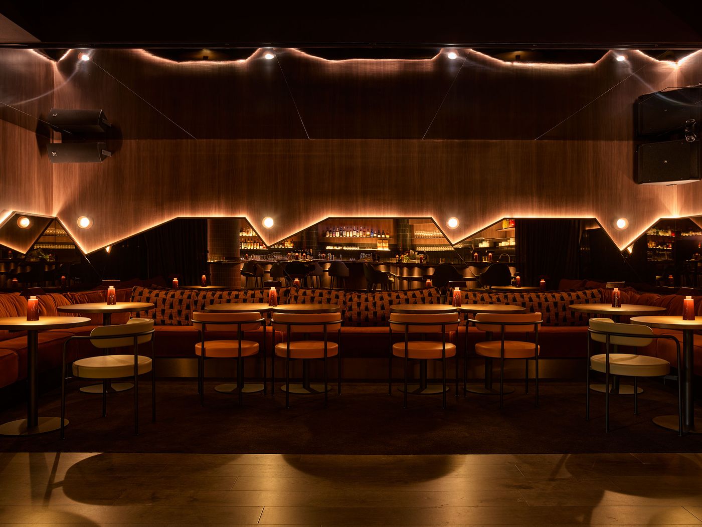 A stylish bar interior with warm lighting, featuring cozy seating, circular tables, and a sleek, modern design.