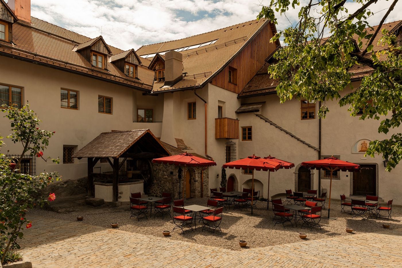 Castel Badia's inner courtyard gathers around a timber-roofed draw well beneath dormered roofs and a jutting wooden balcony. Red scalloped parasols shade wrought-iron chairs on raked gravel, while climbing roses, cobbled paving and a painted Madonna lunette above an arched doorway soften the rendered façades. Castel Badia's inner courtyard gathers around a timber-roofed draw well beneath dormered roofs and a jutting wooden balcony. Red scalloped parasols shade wrought-iron chairs on raked gravel, while climbing roses, cobbled paving and a painted Madonna lunette above an arched doorway soften the rendered façades.