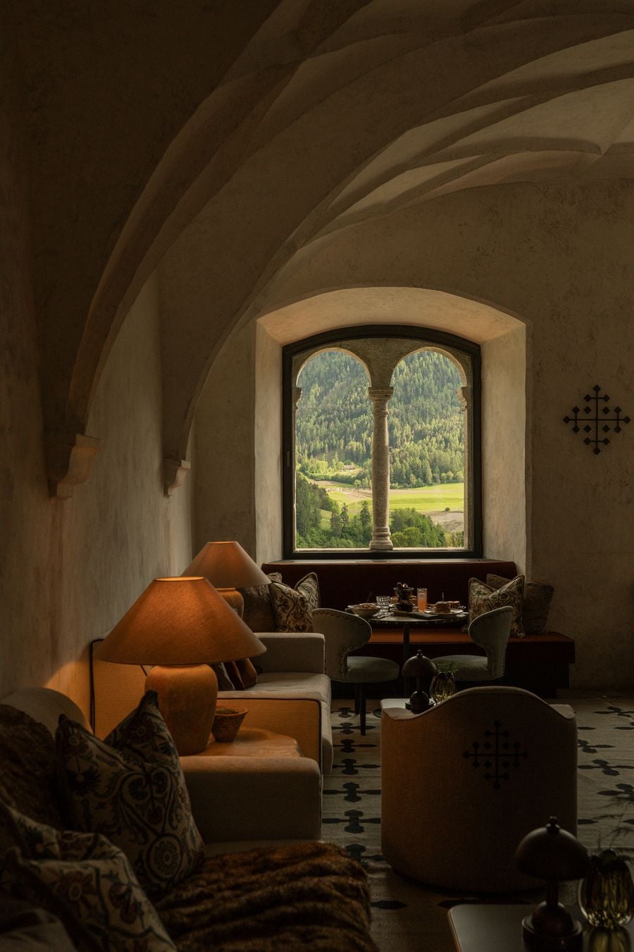 Beneath a sweeping vault in the Abbess's Hall, a twin-arched window with a slender stone column frames a vivid summer view of forest and meadow. Below it, a breakfast table set between studded chairs and a velvet banquette glows in soft lamplight, edged by patterned cushions and the cross-motif rug. Beneath a sweeping vault in the Abbess's Hall, a twin-arched window with a slender stone column frames a vivid summer view of forest and meadow. Below it, a breakfast table set between studded chairs and a velvet banquette glows in soft lamplight, edged by patterned cushions and the cross-motif rug.