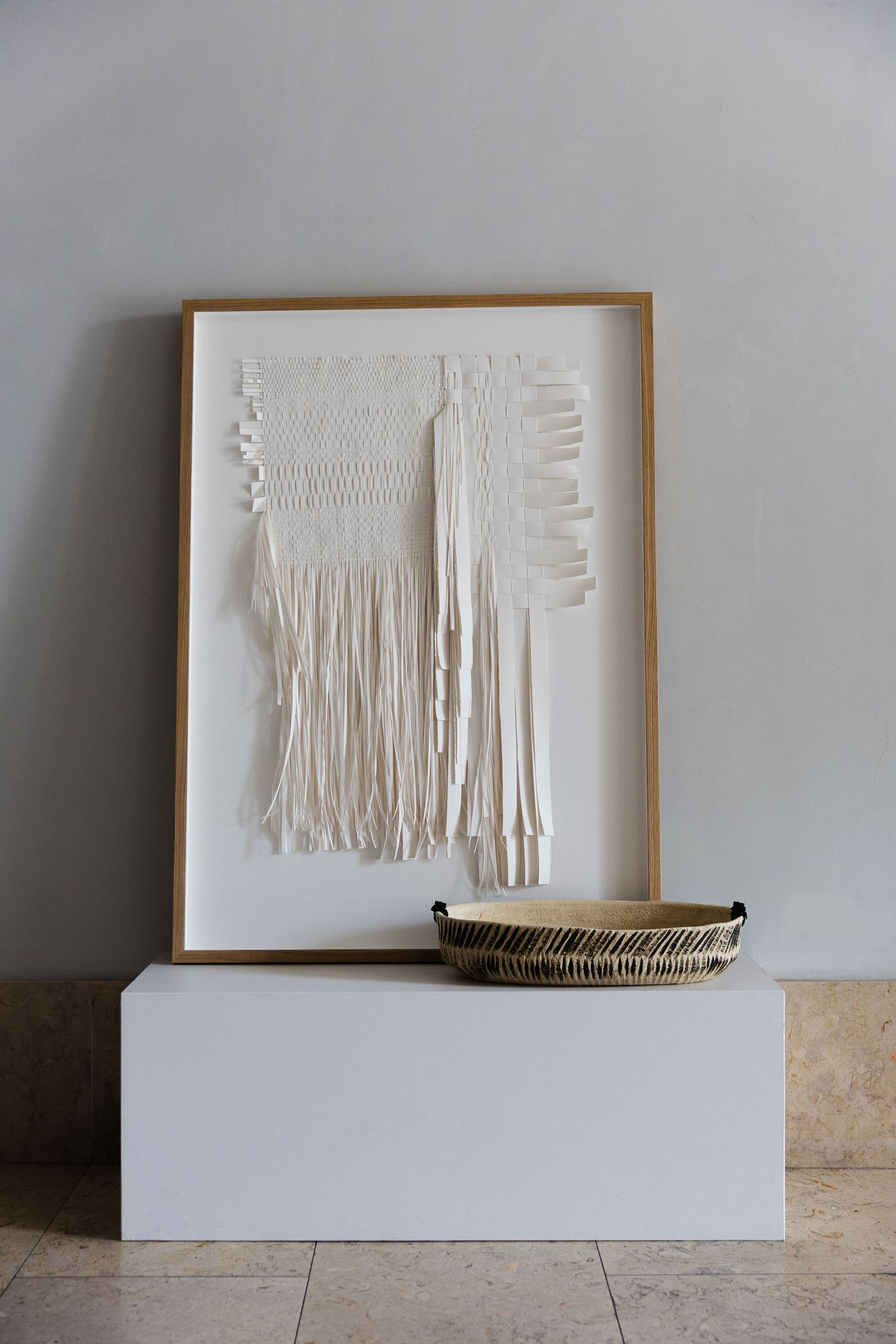 A framed, minimalist textile artwork by Anna Westerlund, featuring layers of cut and woven white paper or fabric strips, stands on a pristine white plinth at Santa Clara 1728. Beside it, a shallow, patterned woven basket adds a contrasting organic texture. The light wood frame and clean composition evoke a serene, Japandi-inspired aesthetic. A framed, minimalist textile artwork by Anna Westerlund, featuring layers of cut and woven white paper or fabric strips, stands on a pristine white plinth at Santa Clara 1728. Beside it, a shallow, patterned woven basket adds a contrasting organic texture. The light wood frame and clean composition evoke a serene, Japandi-inspired aesthetic.