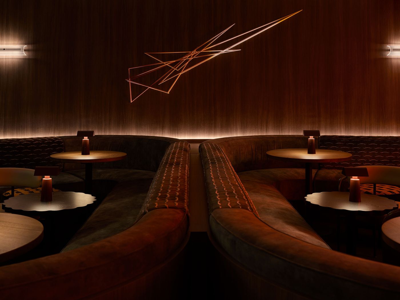 Intimate lounge with curved, plush seating, wooden tables, and ambient lighting, featuring an abstract light installation above. Intimate lounge with curved, plush seating, wooden tables, and ambient lighting, featuring an abstract light installation above.