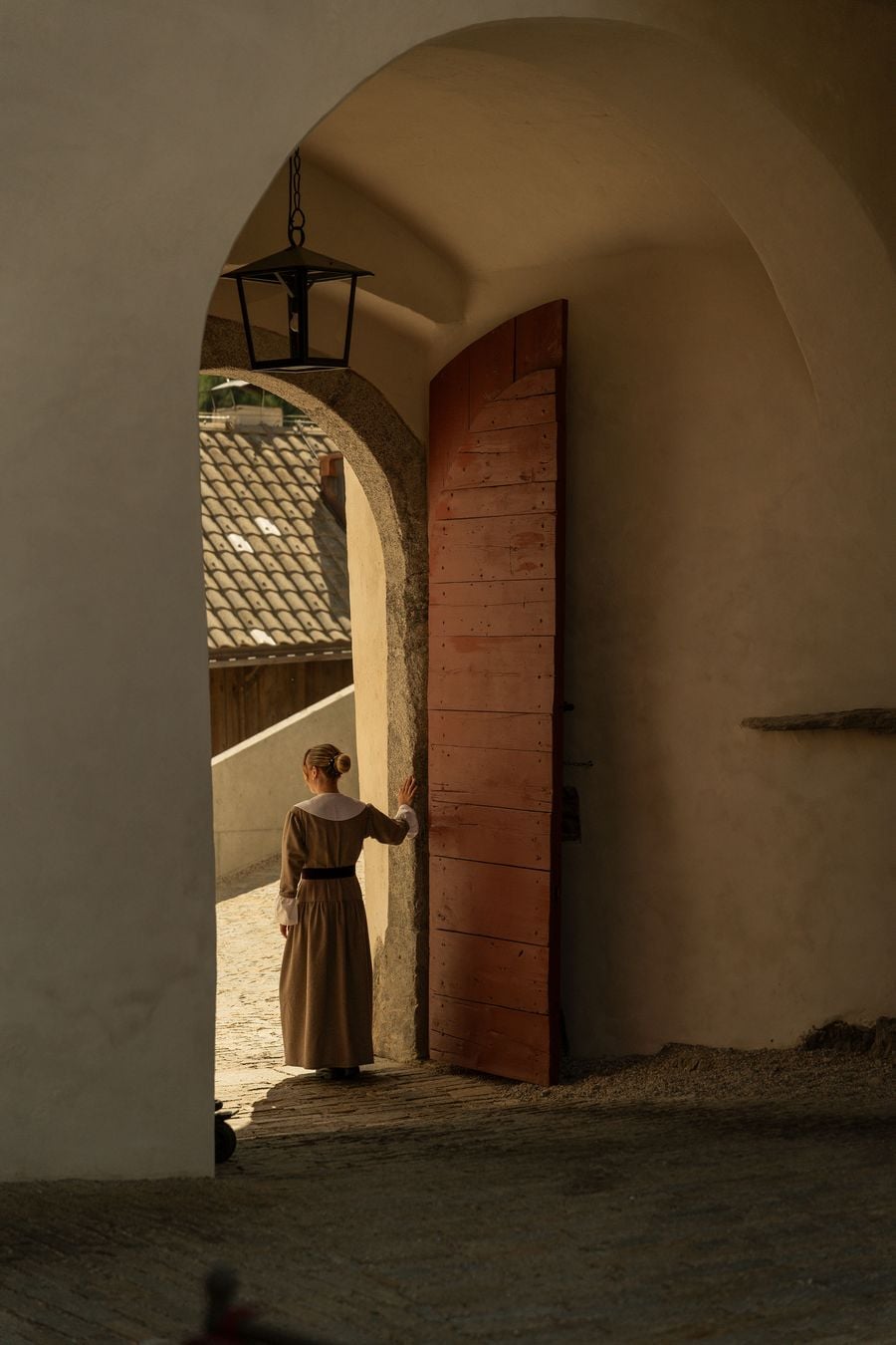 A server in a long wool dress with a white collar rests her hand on a heavy plank door, painted oxblood, in Castel Badia's vaulted gate passage. A wrought-iron lantern hangs from the plastered arch, while the granite-framed opening reveals sunlit cobbles and a stone-tiled roof beyond. A server in a long wool dress with a white collar rests her hand on a heavy plank door, painted oxblood, in Castel Badia's vaulted gate passage. A wrought-iron lantern hangs from the plastered arch, while the granite-framed opening reveals sunlit cobbles and a stone-tiled roof beyond.