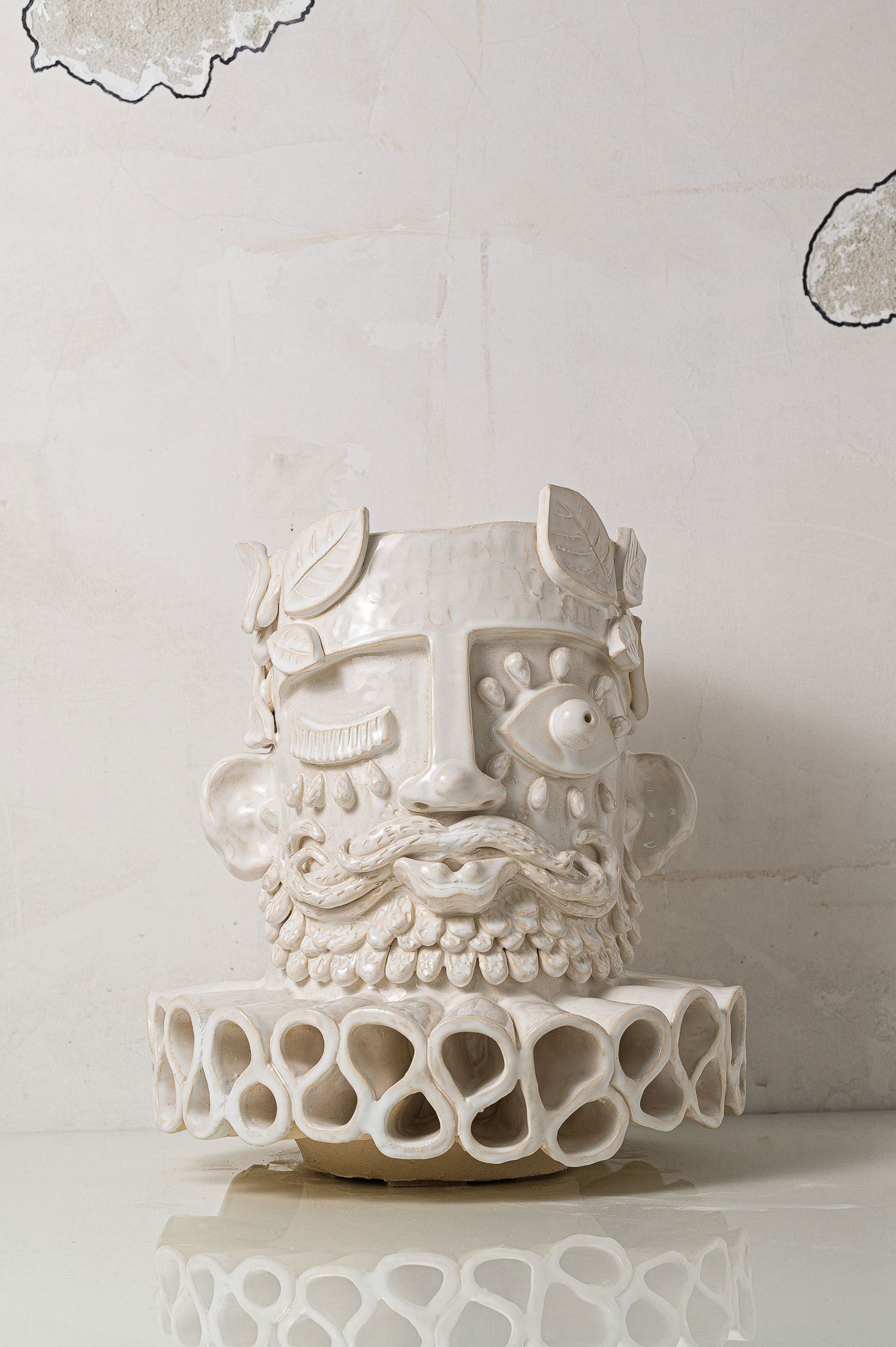 A highly textured, off-white ceramic sculpture by Carlos Manuel Gonçalves features a stylized human face with closed eyes, a prominent nose, and an elaborate beard. Below the head, a wide, ruffled collar with intricate loop patterns forms the base. The raw, almost unfinished aesthetic, set against a subtly textured white wall, gives the piece an ancient, artisanal quality. A highly textured, off-white ceramic sculpture by Carlos Manuel Gonçalves features a stylized human face with closed eyes, a prominent nose, and an elaborate beard. Below the head, a wide, ruffled collar with intricate loop patterns forms the base. The raw, almost unfinished aesthetic, set against a subtly textured white wall, gives the piece an ancient, artisanal quality.
