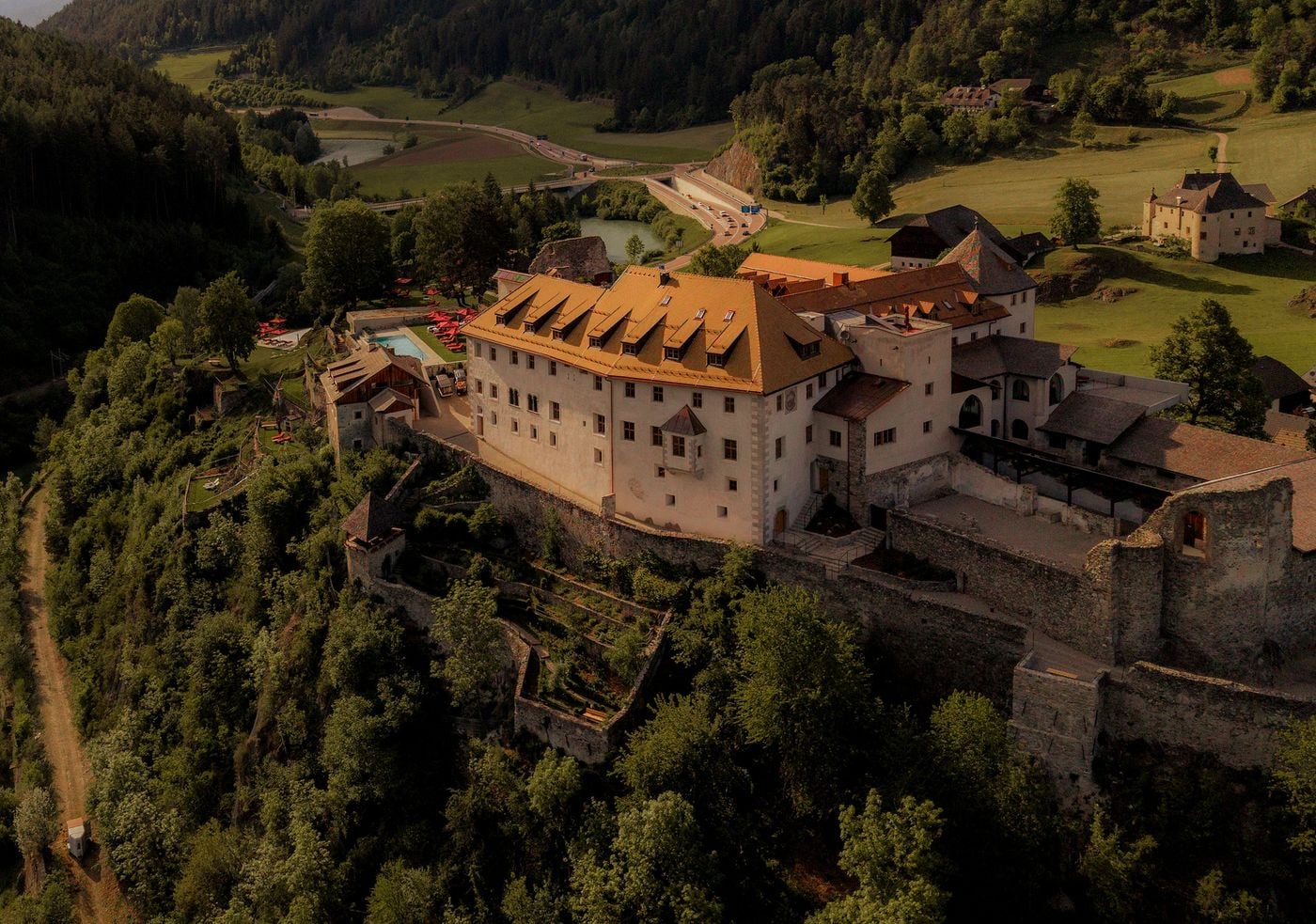 Late sun warms Castel Badia's main wing, its tiled roof lined with dormers and its façade marked by an oriel window and painted quoins. Medieval curtain walls, crumbling ruins and a walled terrace garden step down the wooded slope, while the pool, red parasols, river and valley road lie beyond. Late sun warms Castel Badia's main wing, its tiled roof lined with dormers and its façade marked by an oriel window and painted quoins. Medieval curtain walls, crumbling ruins and a walled terrace garden step down the wooded slope, while the pool, red parasols, river and valley road lie beyond.