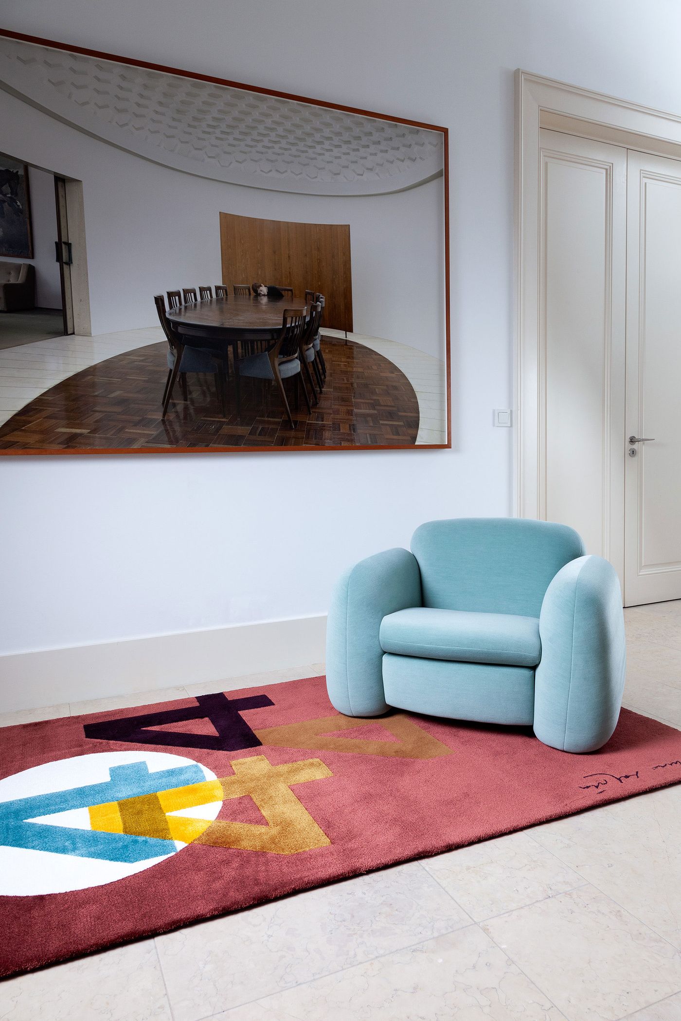 A spacious room features a vibrant red rug with bold, abstract shapes and letters in blue, yellow, and white, designed by Atelier Daciano da Costa. A plush, light blue armchair rests on the rug, complemented by a large framed photograph of a modern, circular dining room on the wall. The contrasting elements create an inviting yet sophisticated interior. A spacious room features a vibrant red rug with bold, abstract shapes and letters in blue, yellow, and white, designed by Atelier Daciano da Costa. A plush, light blue armchair rests on the rug, complemented by a large framed photograph of a modern, circular dining room on the wall. The contrasting elements create an inviting yet sophisticated interior.
