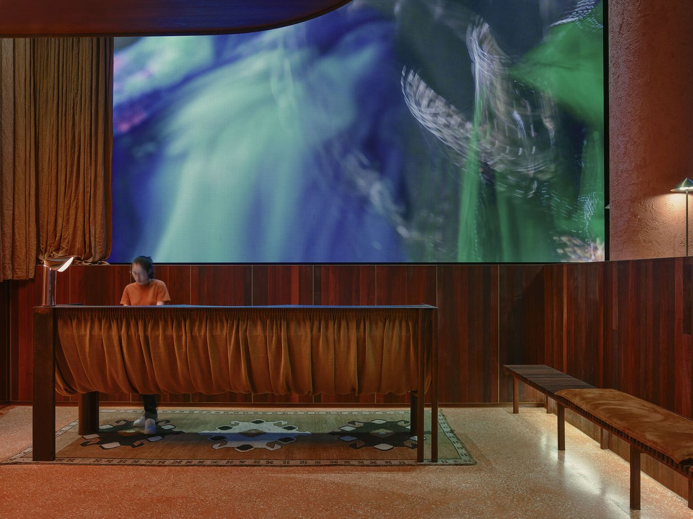 An intimate space featuring a dark wood counter and a large screen displaying an abstract colorful visual, with dim lighting and cozy seating. An intimate space featuring a dark wood counter and a large screen displaying an abstract colorful visual, with dim lighting and cozy seating.