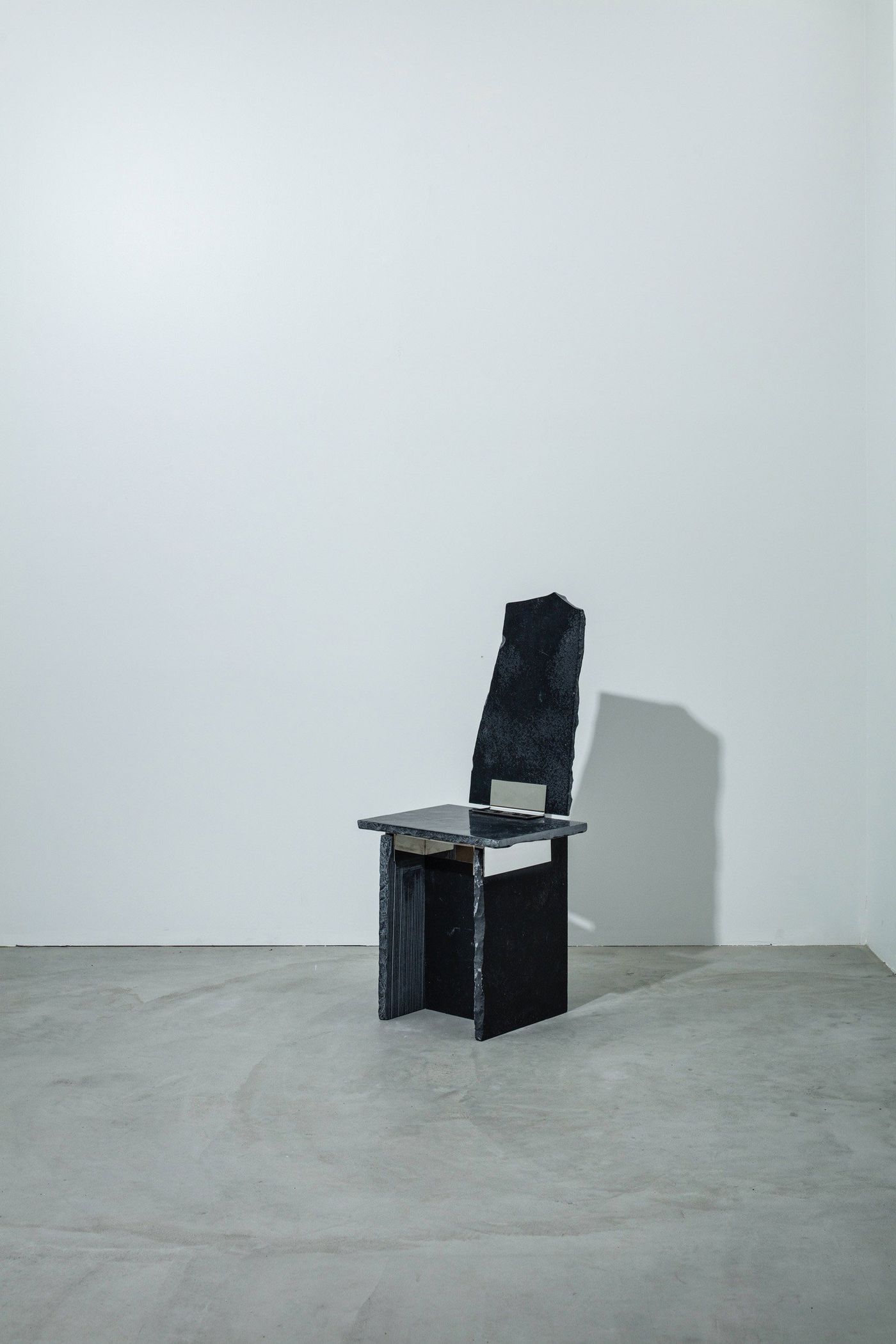 A minimalist black stone chair with an irregular backrest and a slab seat, set against a plain white wall.